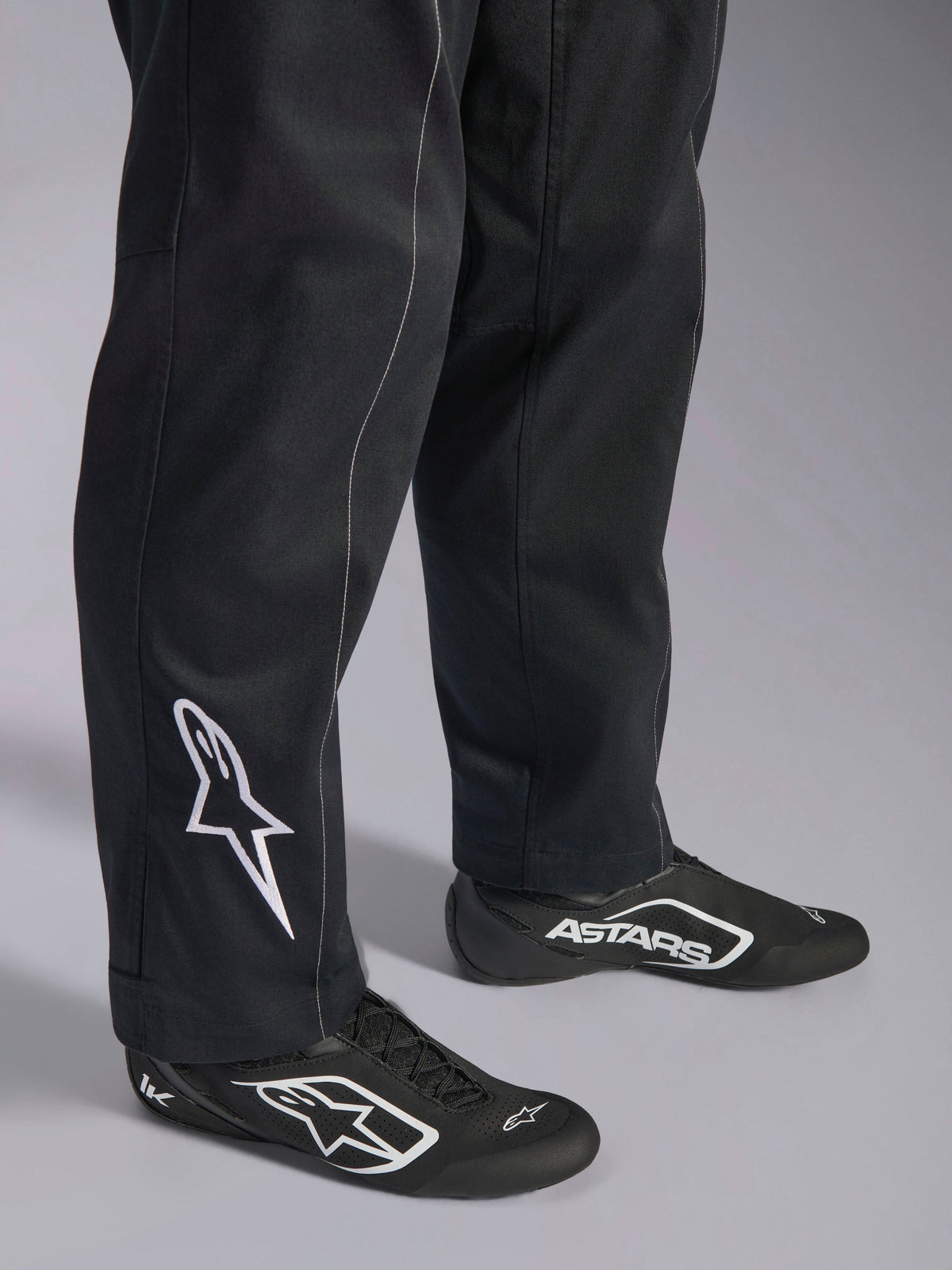 Fato Alpinestars Kart Indoor V2, fato de karting preto, detalhe da parte inferior das pernas e dos pés com o logótipo Alpinestars branco na perna, usado com calçado de competição preto e branco a condizer, concebido para karting recreativo.