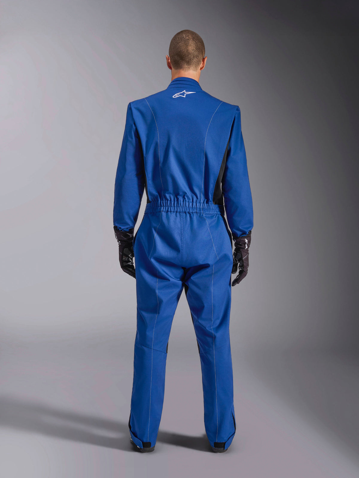 Fato Alpinestars Kart Indoor V2, azul, vista traseira, fato de karting de uma peça com logótipo branco na parte superior das costas, cintura elástica e detalhes pretos, usado com luvas de corrida pretas