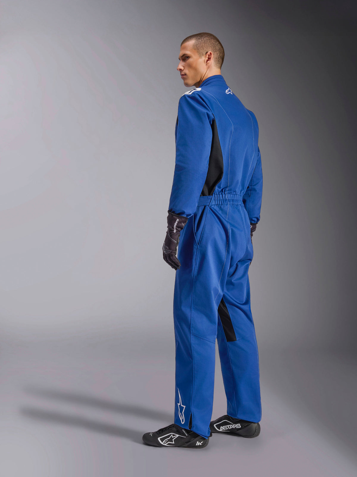 Alpinestars Kart Indoor V2 Suit, fato de karting azul, vista lateral e traseira, fato de competição de peça única com painéis laterais pretos e detalhes no interior da perna, cintura elástica e logótipos brancos, usado com luvas e calçado de competição pretos