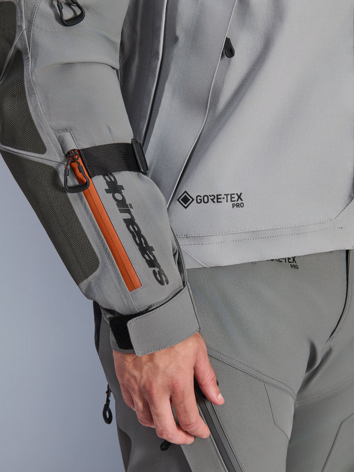 Casaco Alpinestars Nazca 3L GORE-TEX PRO, casaco de aventura em cinzento escuro e cinzento médio, detalhe em grande plano da manga com marca preta e detalhe de fecho cor-de-laranja, logótipo GORE-TEX PRO no tronco, construção têxtil durável com reforço no cotovelo