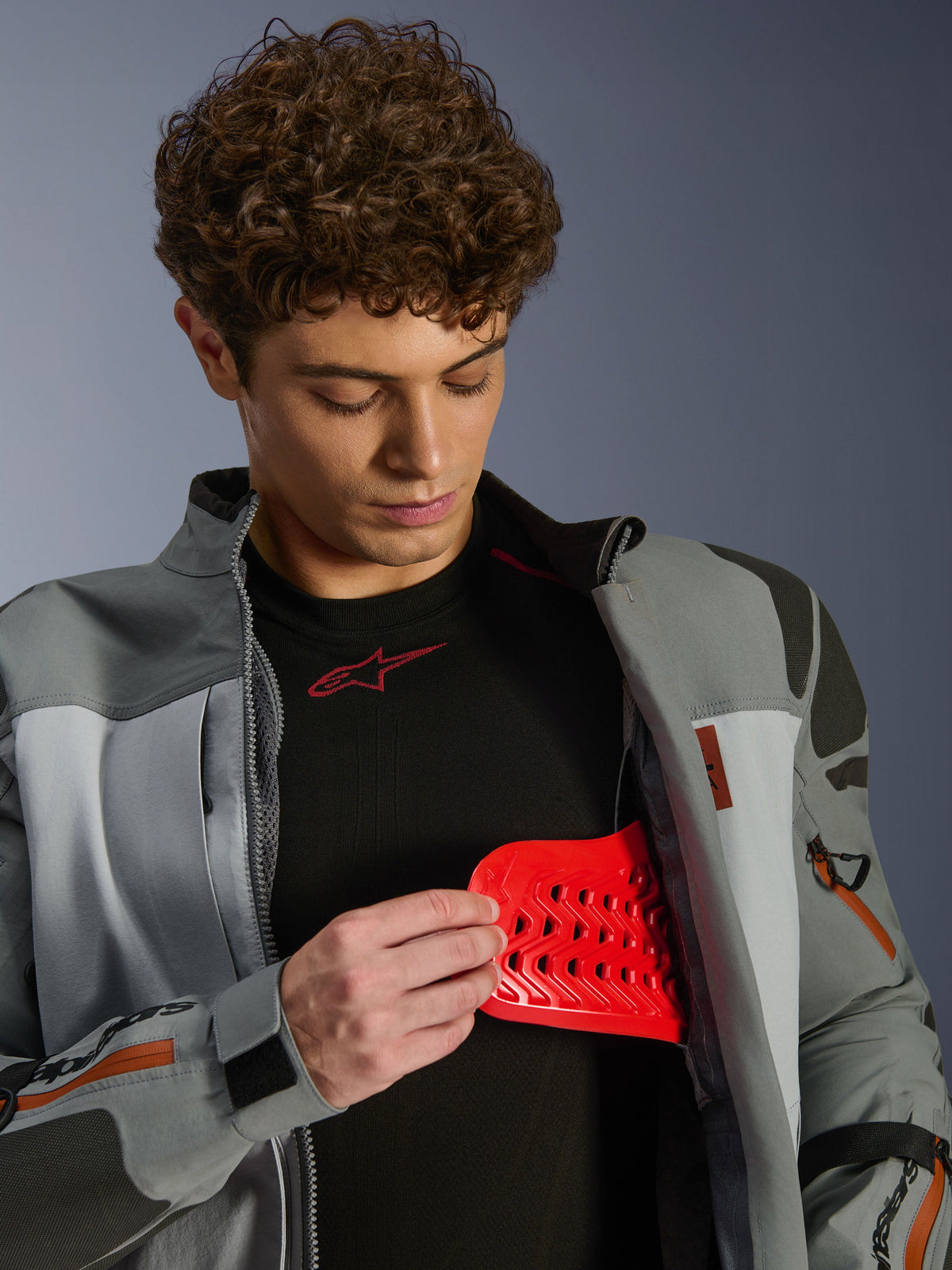 Modelo masculino com casaco Alpinestars Nazca 3L GORE-TEX PRO, cinzento escuro e cinzento médio, a mostrar a proteção Nucleon PLASMA Pro vermelha, vestuário de condução de aventura, casaco de motociclismo premium com detalhes técnicos