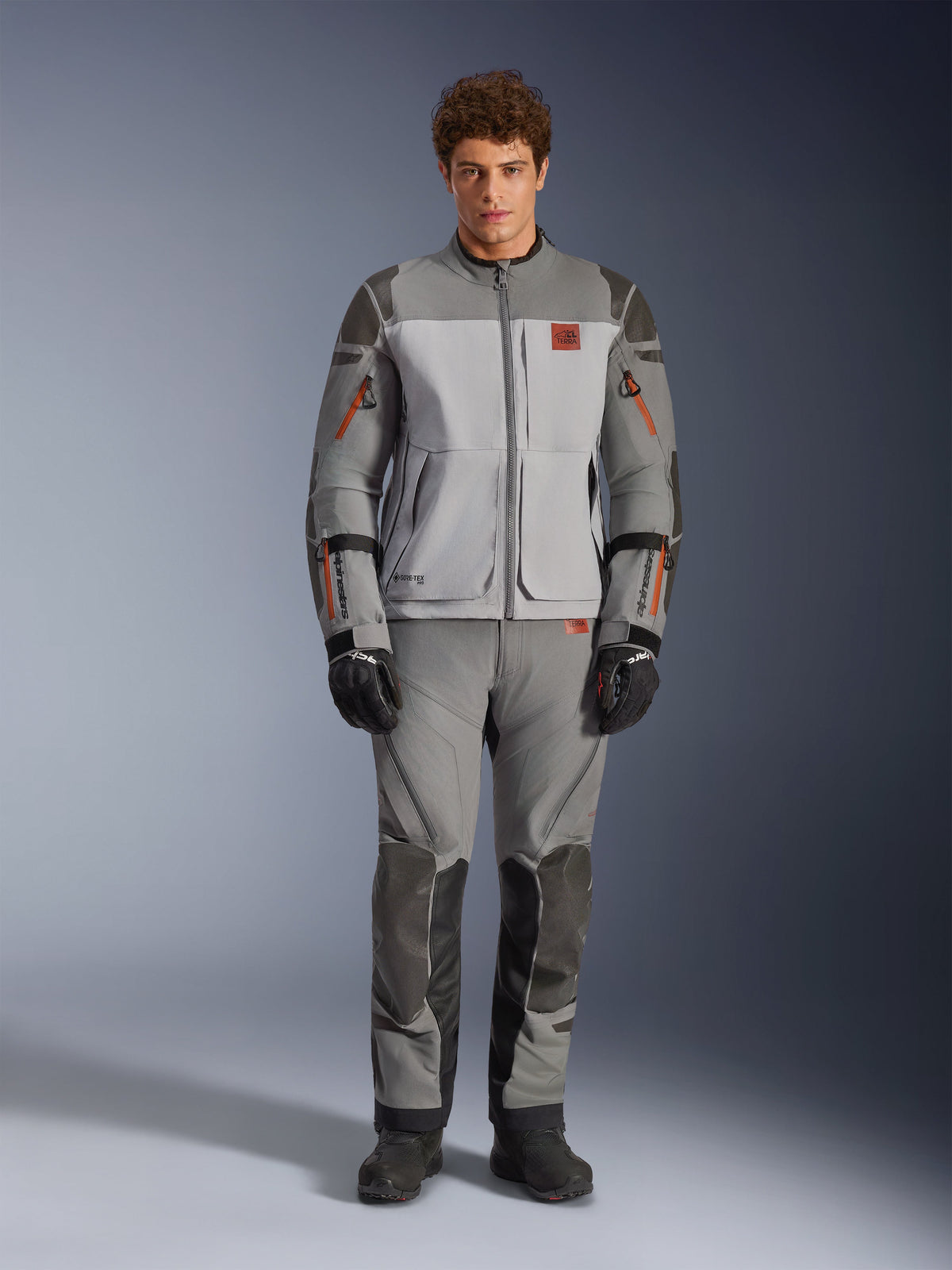 Alpinestars Nazca 3L GORE-TEX PRO Jacket, casaco de motociclismo de aventura, cinzento escuro e cinzento médio, usado por um modelo com calças a condizer e luvas pretas, com detalhes de fecho cor de laranja e logótipo vermelho no peito, equipamento de proteção técnica para condução extrema