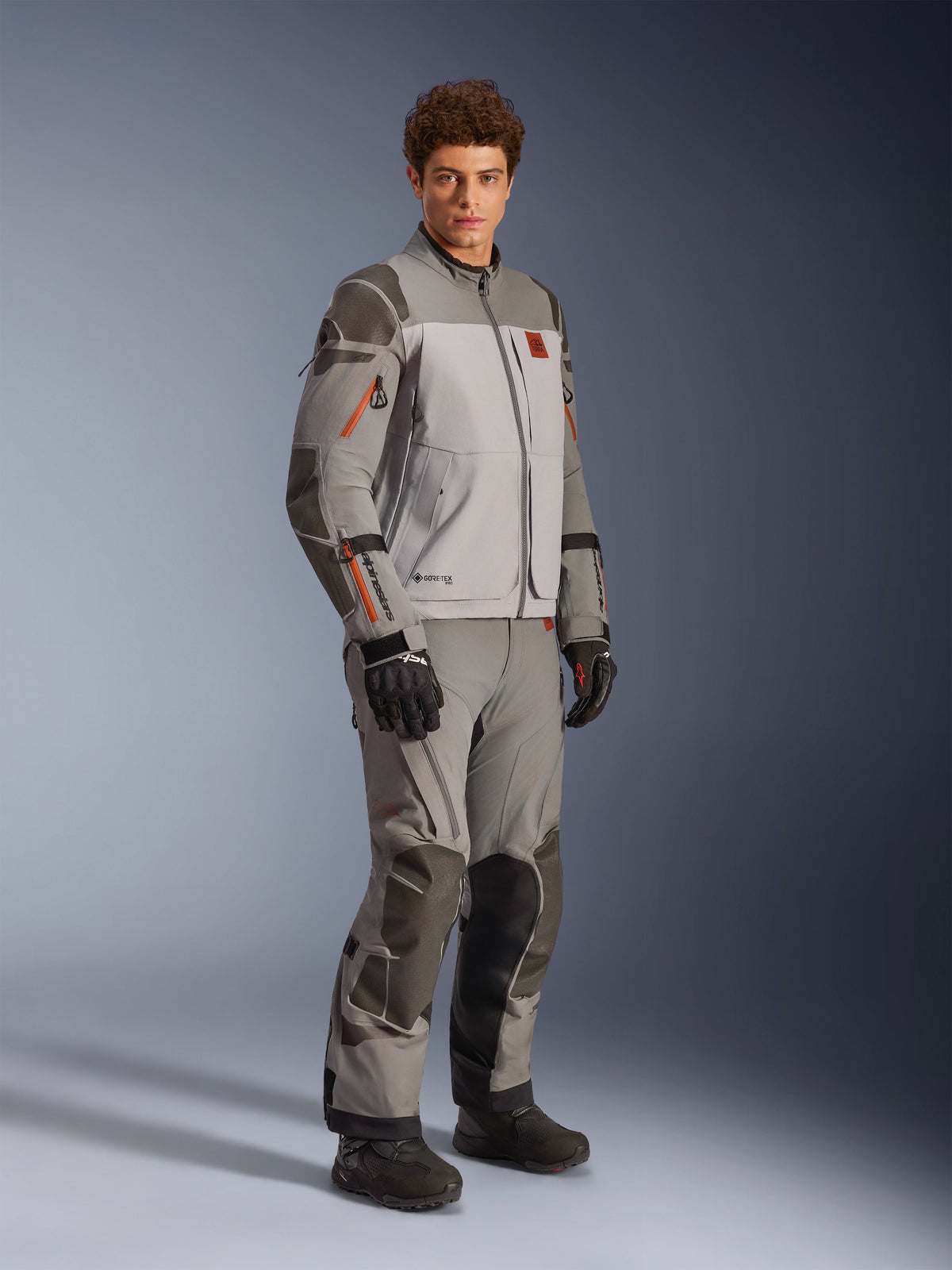 Casaco Alpinestars Nazca 3L GORE-TEX PRO em cinzento escuro e cinzento médio, conjunto de motociclismo de aventura com calças a condizer e luvas pretas, com detalhes de fechos cor-de-laranja, reforços protetores Superfabric® e logótipo GORE-TEX.