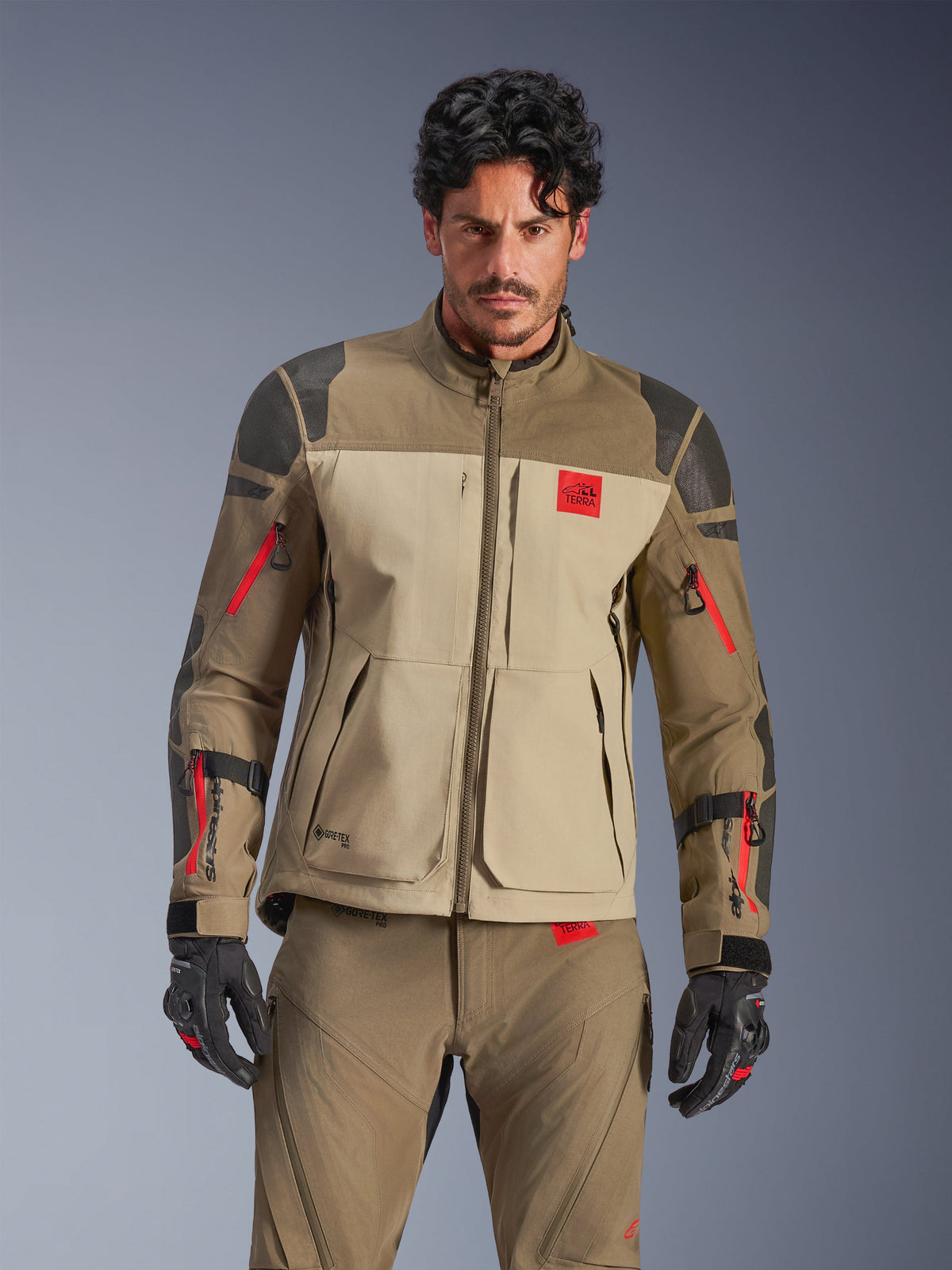 Alpinestars Nazca 3L GORE-TEX PRO Jacket, adventure motorcycle touring jacket, Walnut Rock Brown and beige with red accents, featuring black Superfabric shoulder reinforcements and red sleeve zippers, worn by rider with matching gear, premium technical adventure outerwear