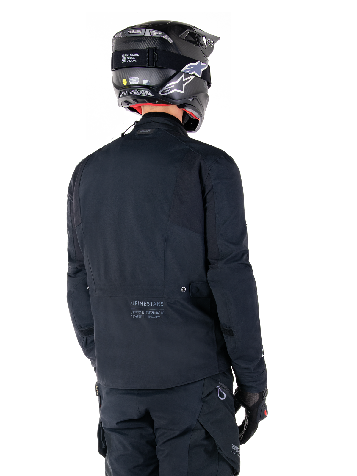 Casaco Alpinestars St-7 Prime GORE-TEX, preto, vista traseira de um motociclista a usar o casaco com capacete e calças a condizer, com o logótipo Alpinestars e coordenadas geográficas na parte inferior das costas