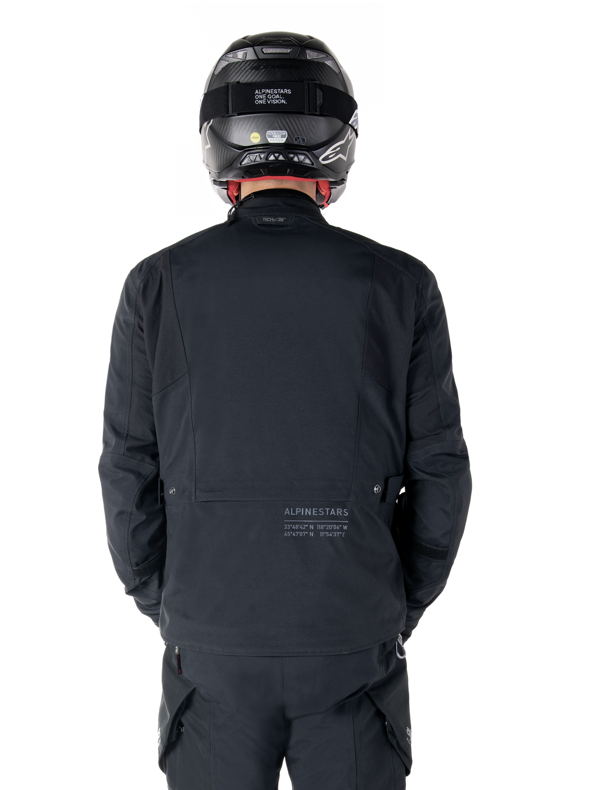 Casaco Alpinestars St-7 Prime GORE-TEX, preto, vista traseira de um motociclista, com logótipo subtil e coordenadas geográficas na parte inferior das costas, usado com capacete e calças pretas a condizer, vestuário de motociclismo elegante e técnico
