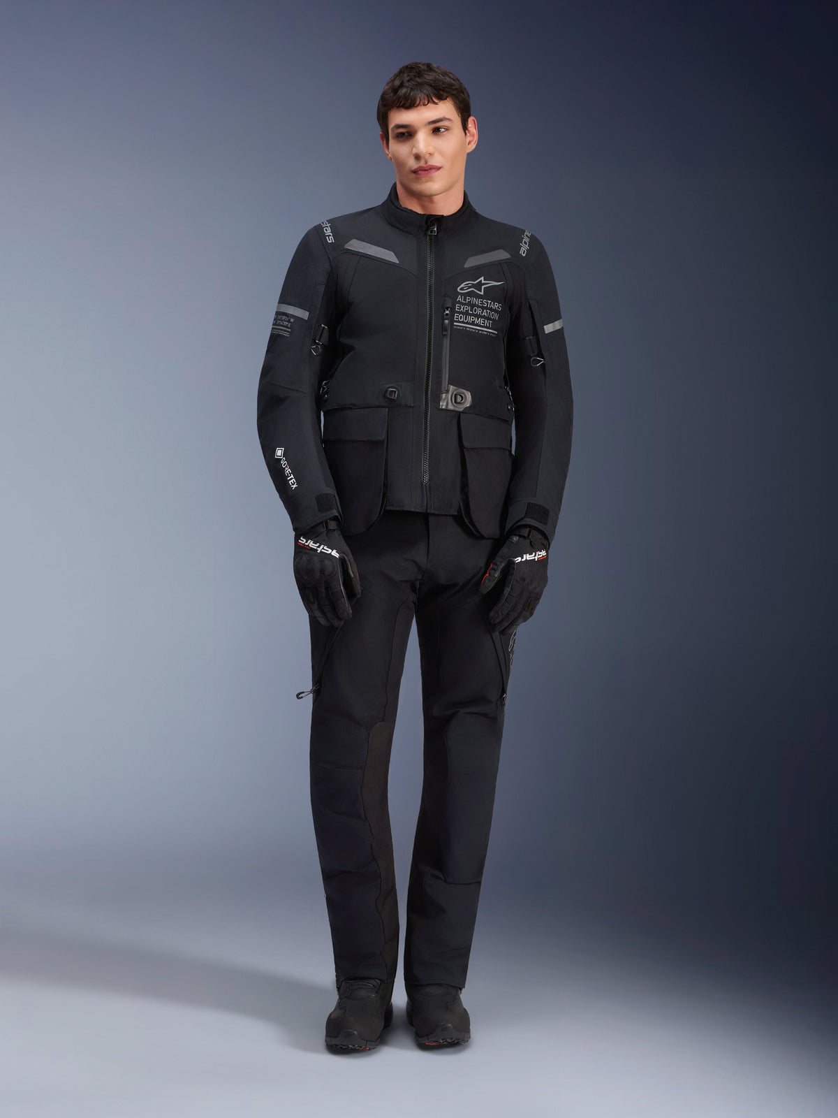 Casaco Alpinestars ST-7 2L GORE-TEX, casaco de motociclismo adventure touring, preto e cinzento escuro, usado com calças e luvas de motociclismo pretas a condizer, com vários bolsos de carga, detalhes refletores e logótipo Alpinestars Exploration Equipment no peito, design funcional para condução em estrada e fora de estrada