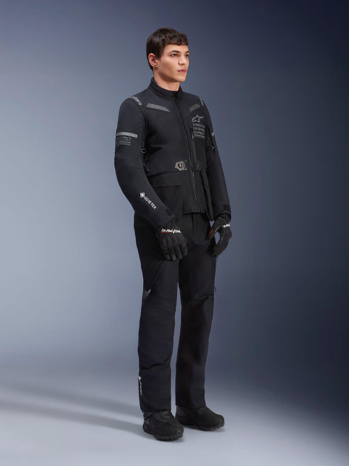 Modelo masculino a usar casaco Alpinestars ST-7 2L GORE-TEX em preto e cinzento escuro, casaco de motociclismo de touring de aventura, calças e luvas pretas a condizer, vista de corpo inteiro a três quartos, com logótipo GORE-TEX e marca Alpinestars Exploration Equipment
