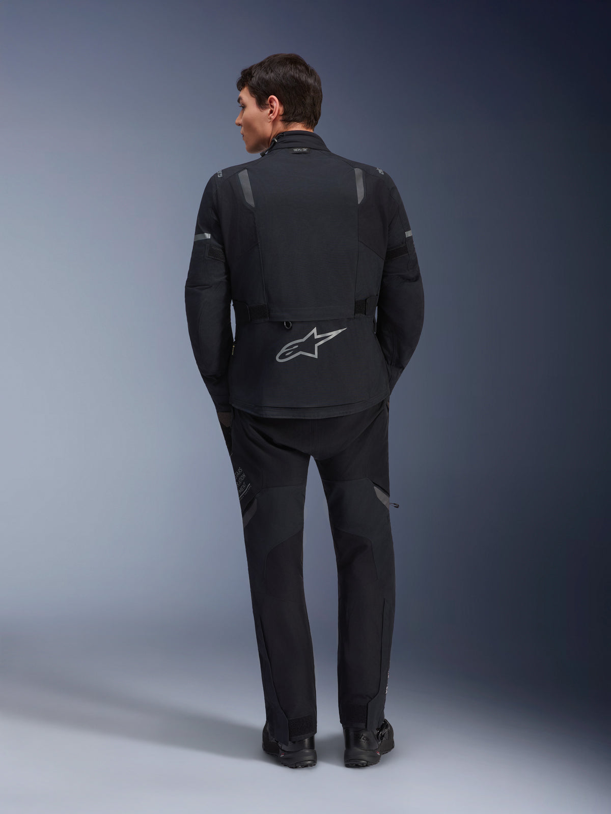 Casaco Alpinestars ST-7 2L GORE-TEX, casaco de motociclismo de aventura, preto e cinzento escuro, vista traseira de um homem a usar o casaco e calças pretas a condizer, com um logótipo Astar refletor grande na parte inferior das costas, equipamento têxtil impermeável para touring e estrada