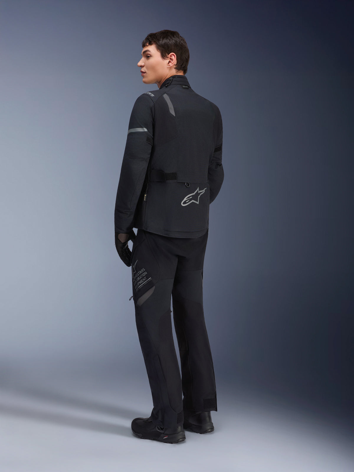 Modelo masculino a usar o Casaco Alpinestars ST-7 2L GORE-TEX em Preto e Cinza Escuro, vista traseira de três quartos mostrando detalhes refletores e logótipo prateado na parte inferior das costas, equipamento de motociclismo adventure touring, combinado com calças de condução e botas pretas.