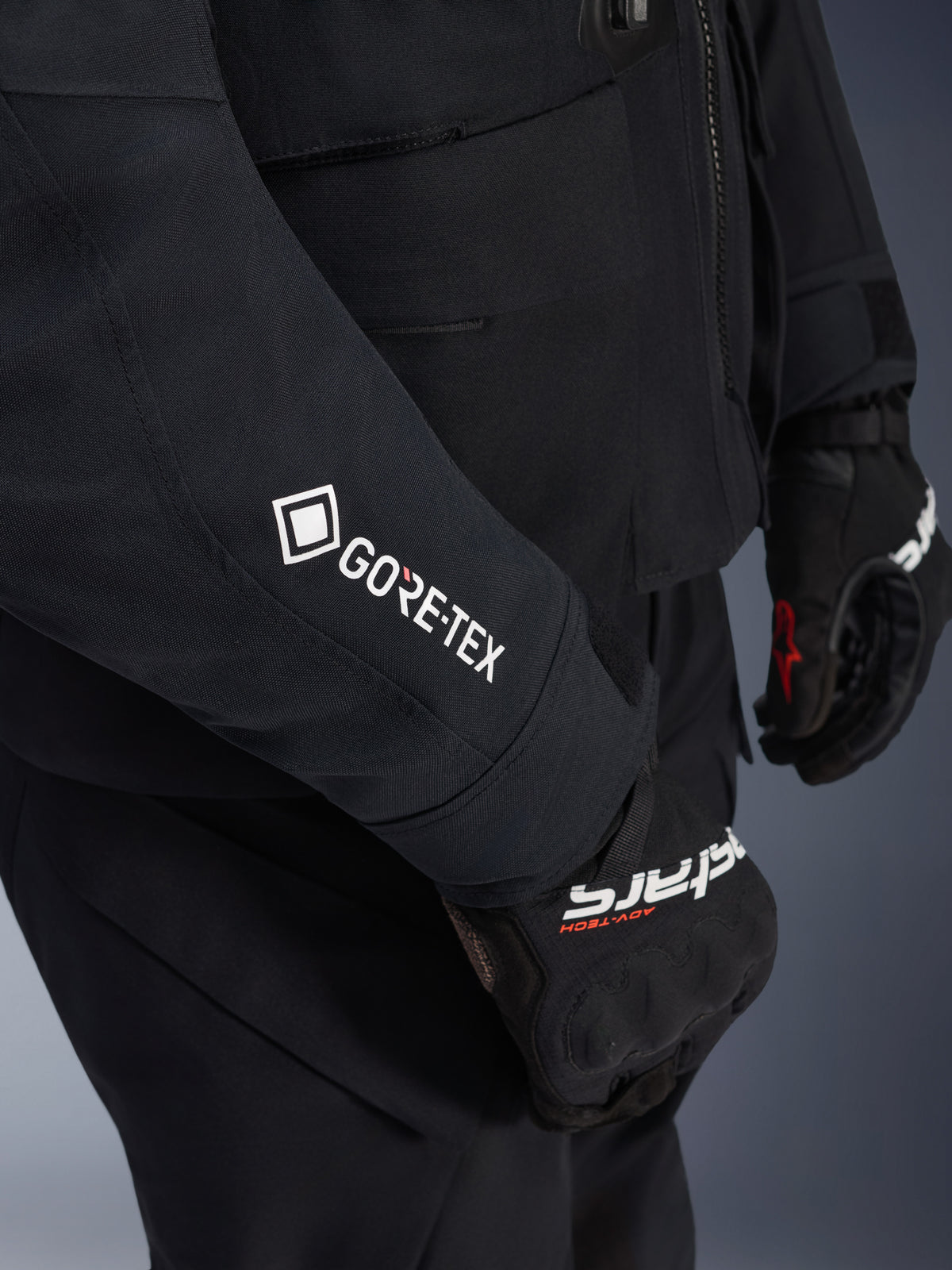 Casaco Alpinestars ST-7 2L GORE-TEX, preto e cinzento escuro, grande plano da manga com logótipo GORE-TEX branco, usado com luvas de aventura pretas, vestuário de motociclismo impermeável