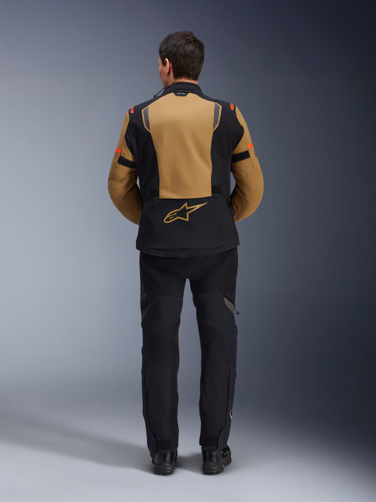 Casaco Alpinestars ST-7 2L GORE-TEX, casaco de motociclismo de aventura, castanho Gobi e preto, vista traseira mostrando painéis ergonómicos e o logótipo Alpinestars, usado com calças e botas de motociclismo pretas, equipamento de touring impermeável
