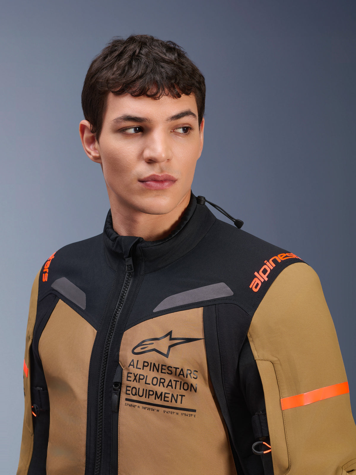Casaco Alpinestars ST-7 2L GORE-TEX, Castanho Gobi e Preto, usado por um modelo masculino num cenário de estúdio, com a marca 'ALPINESTARS EXPLORATION EQUIPMENT' em destaque no peito, logótipos laranja nos ombros e construção técnica em têxtil impermeável para motociclismo de aventura