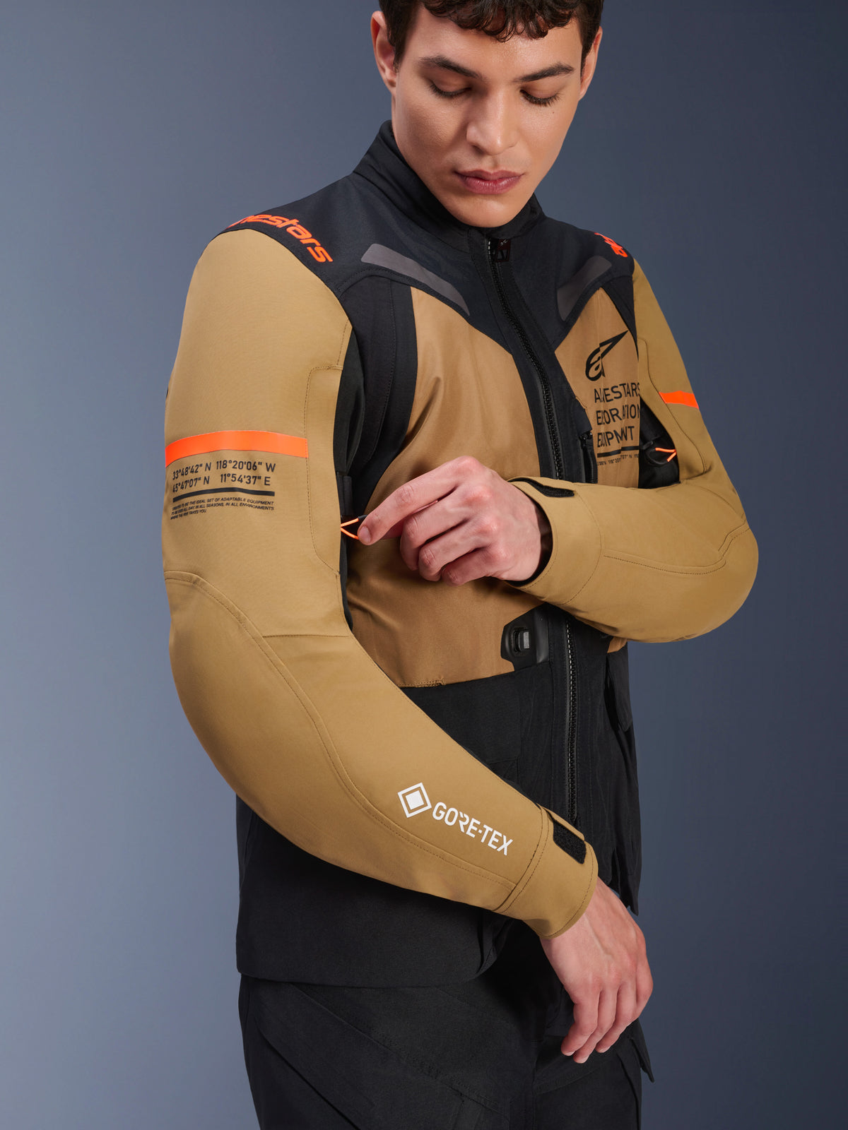 Casaco Alpinestars ST-7 2L GORE-TEX em Castanho Gobi e Preto, casaco de motociclismo adventure-touring vestido por um modelo a ajustar uma abertura de ventilação na manga, com detalhes em laranja, gráficos de coordenadas e logótipo GORE-TEX