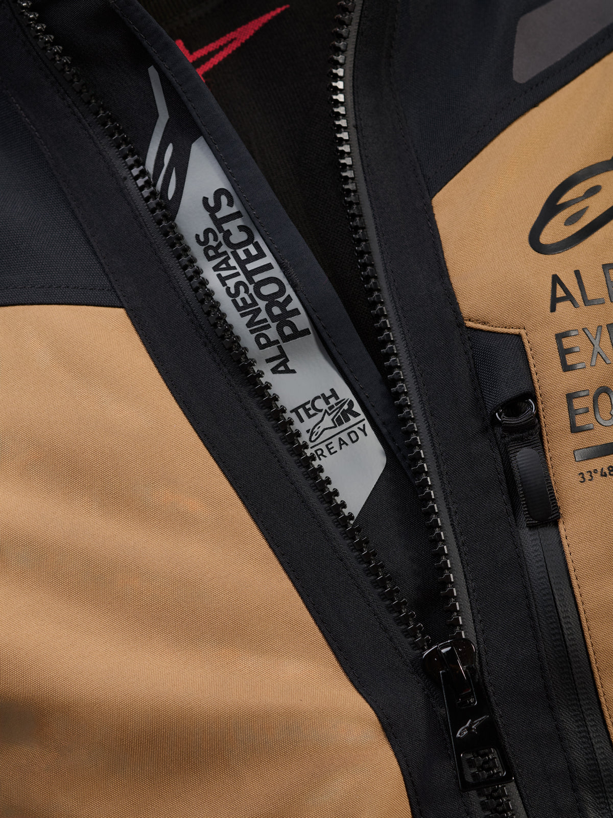 Pormenor do Casaco Alpinestars ST-7 2L GORE-TEX em Castanho Gobi e Preto, apresentando as marcas 'ALPINESTARS PROTECTS' e 'TECH-AIR READY' no interior do fecho principal, com construção têxtil resistente e logótipos no peito.