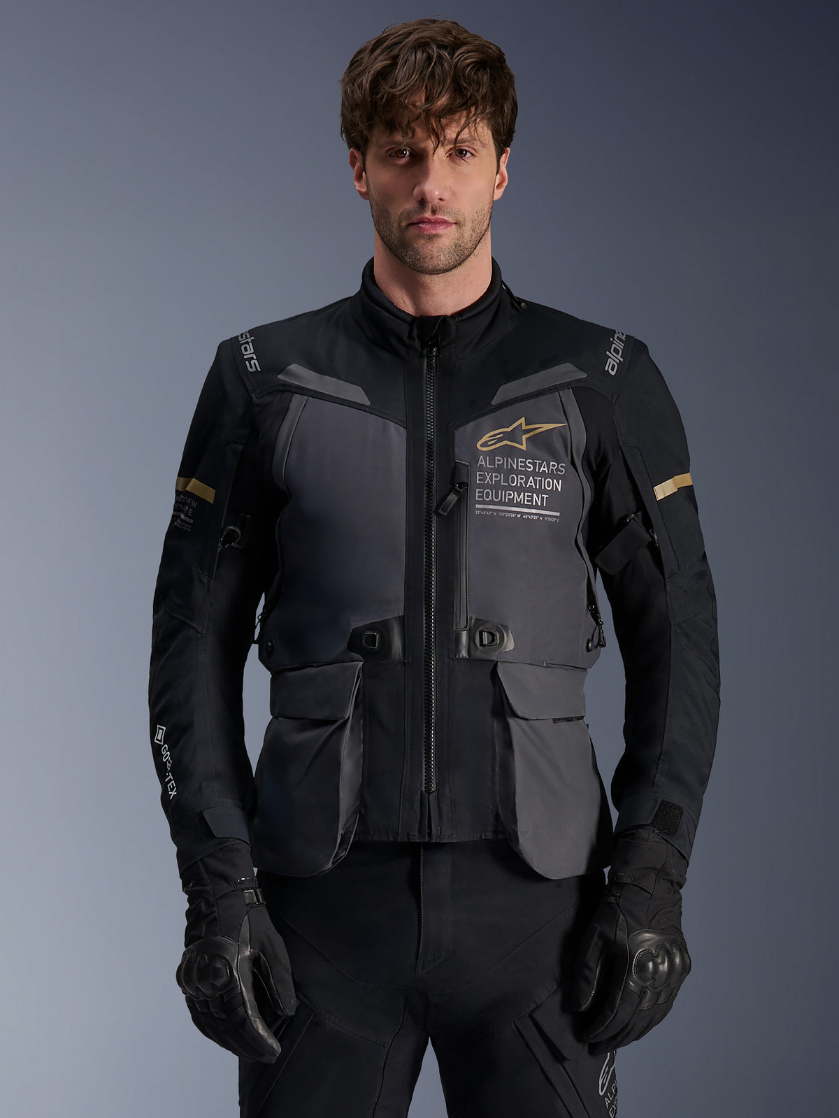 Casaco Alpinestars ST-7 2L GORE-TEX, casaco de motociclismo adventure touring para homem, cinzento escuro e preto com detalhes em caqui, com a marca 'Alpinestars Exploration Equipment', bolsos de carga impermeáveis e gola alta, usado por um modelo com luvas pretas