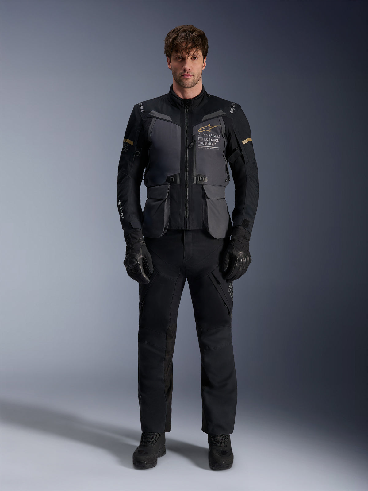 Modelo masculino a usar o Casaco Alpinestars ST-7 2L GORE-TEX em cinzento escuro, preto e caqui escuro, com a inscrição 'ALPINESTARS EXPLORATION EQUIPMENT' no peito, combinado com calças de condução, luvas e botas pretas, vista frontal em estúdio.