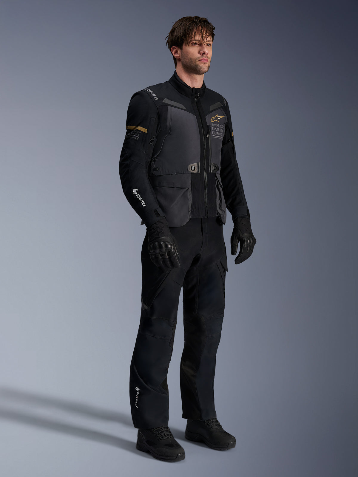 Modelo masculino a usar o Casaco Alpinestars ST-7 2L GORE-TEX em cinzento escuro, preto e caqui escuro, combinado com calças e luvas de aventura pretas a condizer, com camada laminada impermeável, bolsos cargo e logótipos técnicos, ideal para touring de aventura.