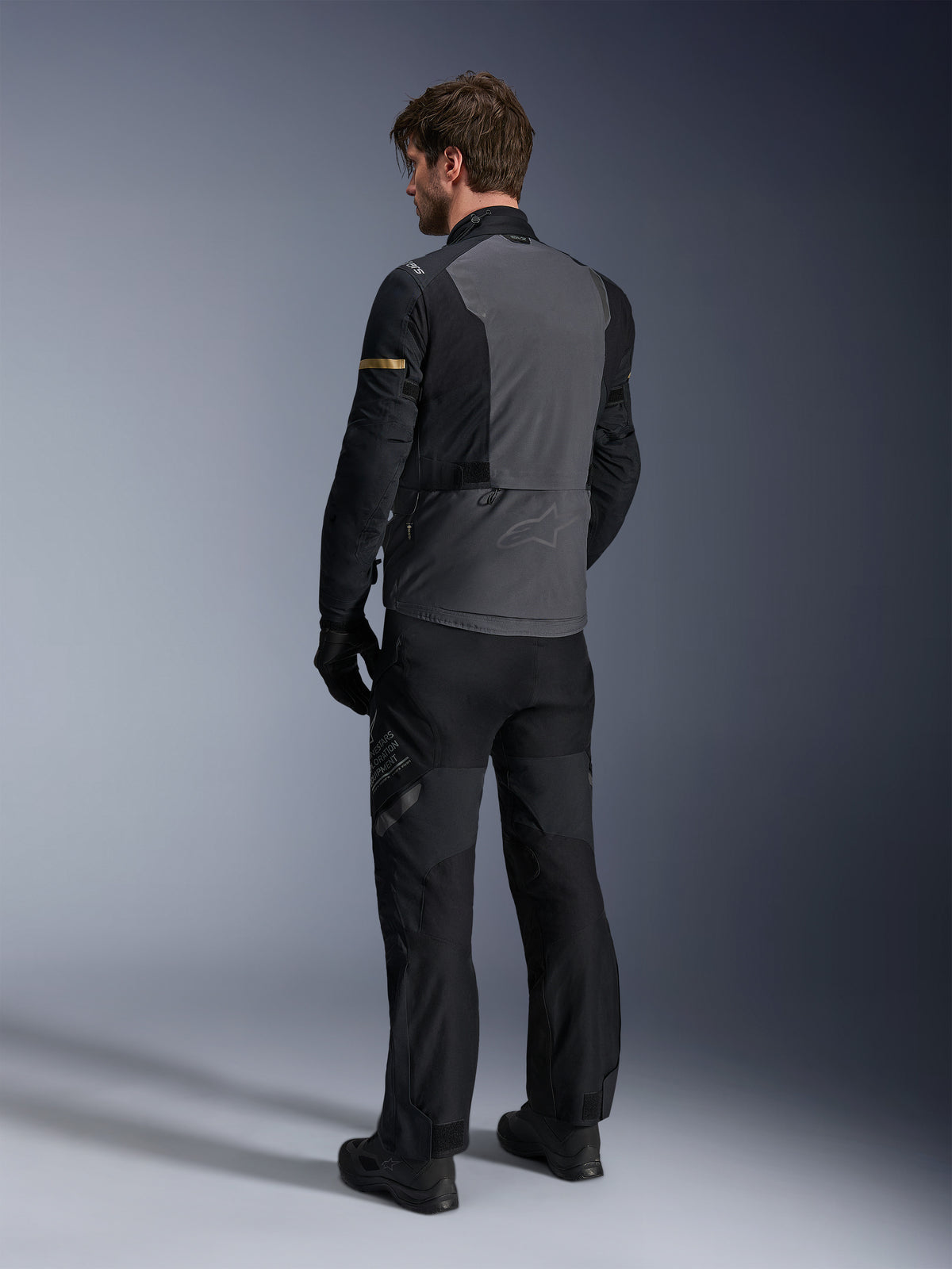 Casaco Alpinestars ST-7 2L GORE-TEX, cinzento escuro e preto com detalhes em Dark Khaki, vista traseira de um motociclista a usar o casaco e calças de motociclismo a condizer, equipamento de touring de aventura com construção laminada impermeável