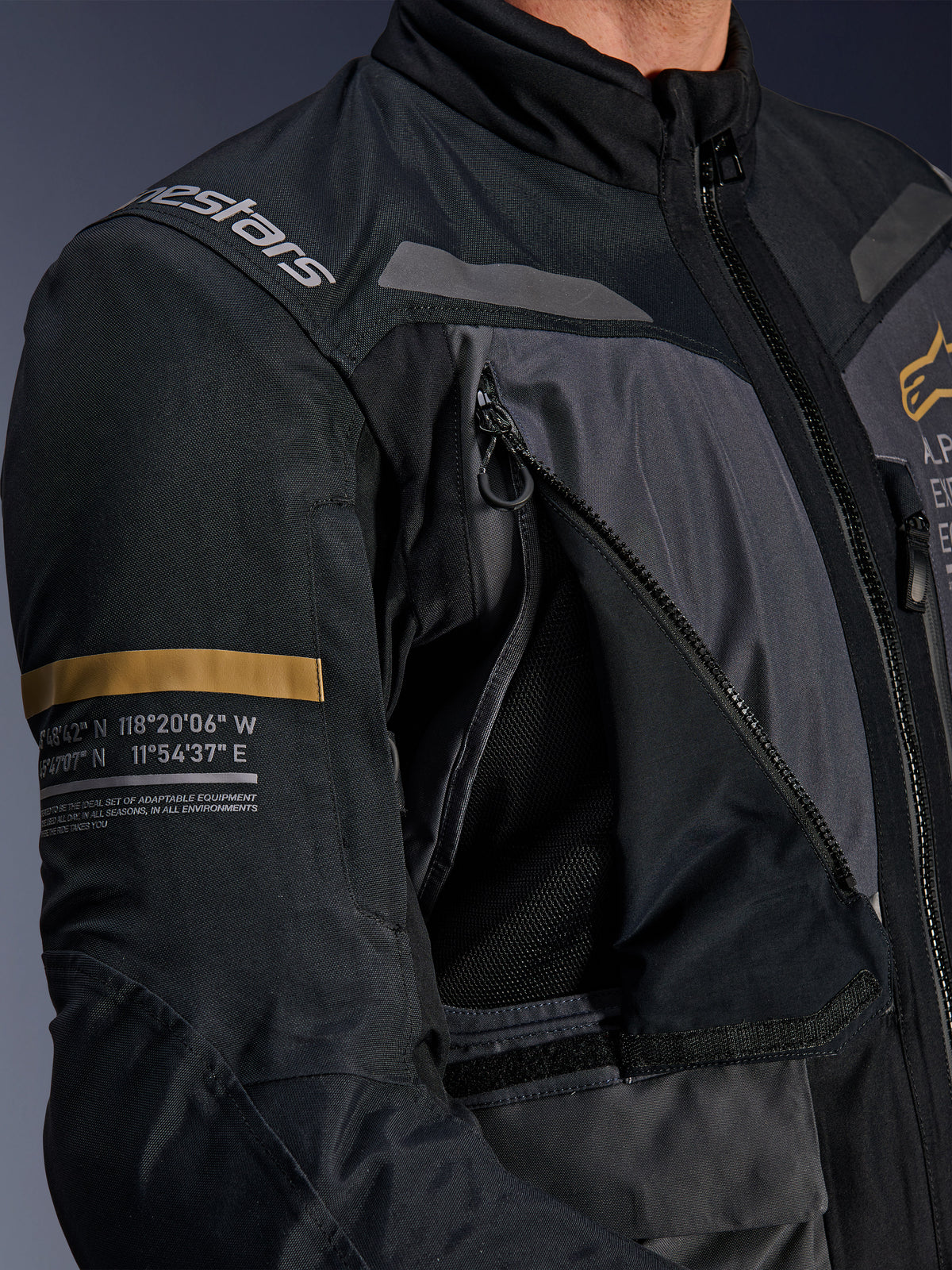 Casaco Alpinestars ST-7 2L GORE-TEX, cinzento escuro e preto, detalhe do painel de ventilação no peito com fecho aberto e malha, logótipo Alpinestars no ombro e risca caqui na manga com coordenadas geográficas impressas, equipamento técnico de motociclismo adventure-touring