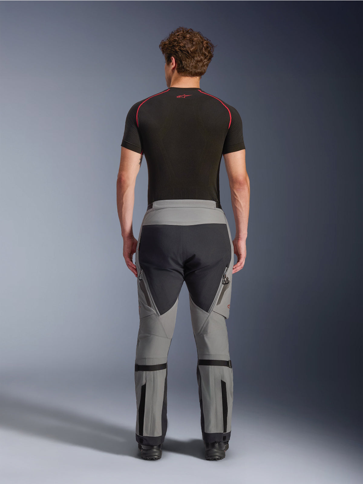 Alpinestars Nazca 3L GORE-TEX PRO Pants, Dark Gray Black adventure motorcycle pants, rear view on a model, technical multi-panel design with gray and black fabric, featuring calf volume adjusters and worn with a black base layer with red accents.