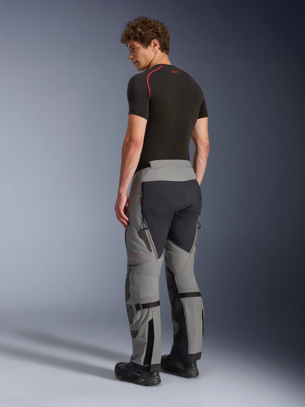 Alpinestars Nazca 3L GORE-TEX PRO Pants, Adventure Riding Pants, Dark Gray and Black, back view of model wearing a black base layer and boots, showing technical panels, ventilation zippers, and calf volume adjusters.