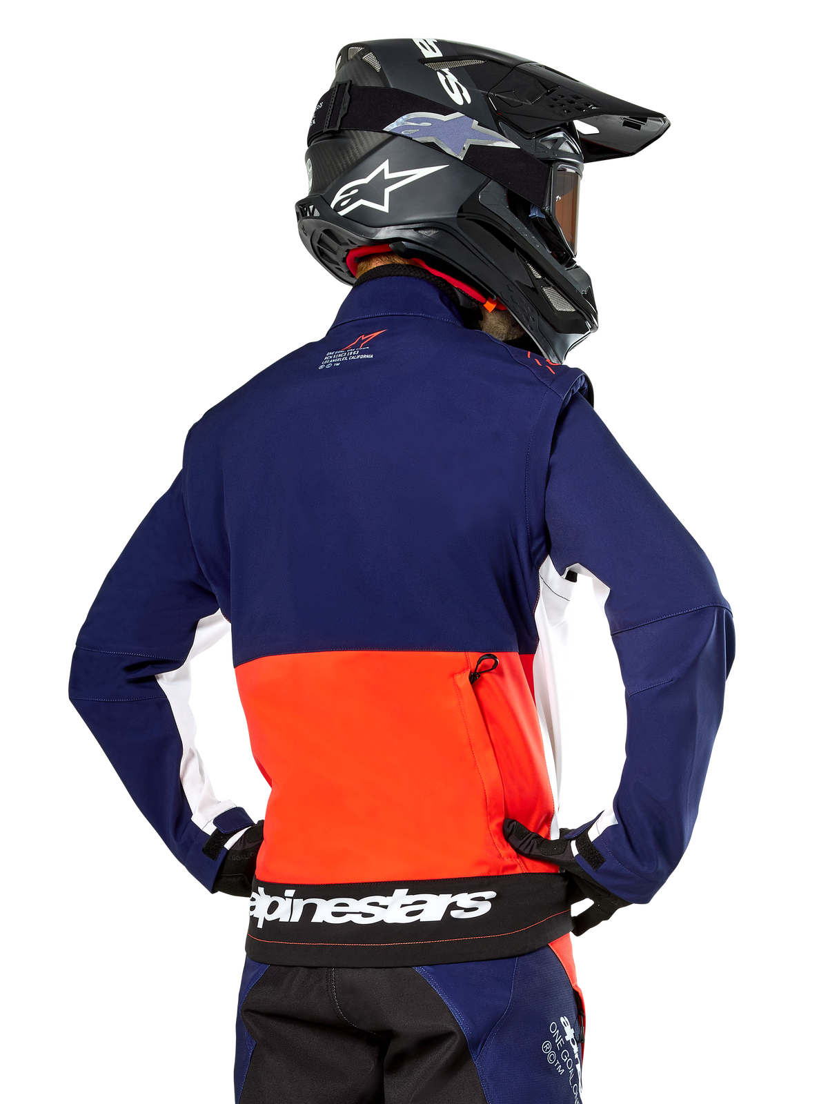 Casaco Alpinestars Lite-Dura Softshell, azul marinho, laranja vibrante e branco, vista traseira usado por um piloto de motocross, com um logótipo branco grande na bainha traseira e bolso de armazenamento com fecho, parte de um equipamento completo de corrida off-road.