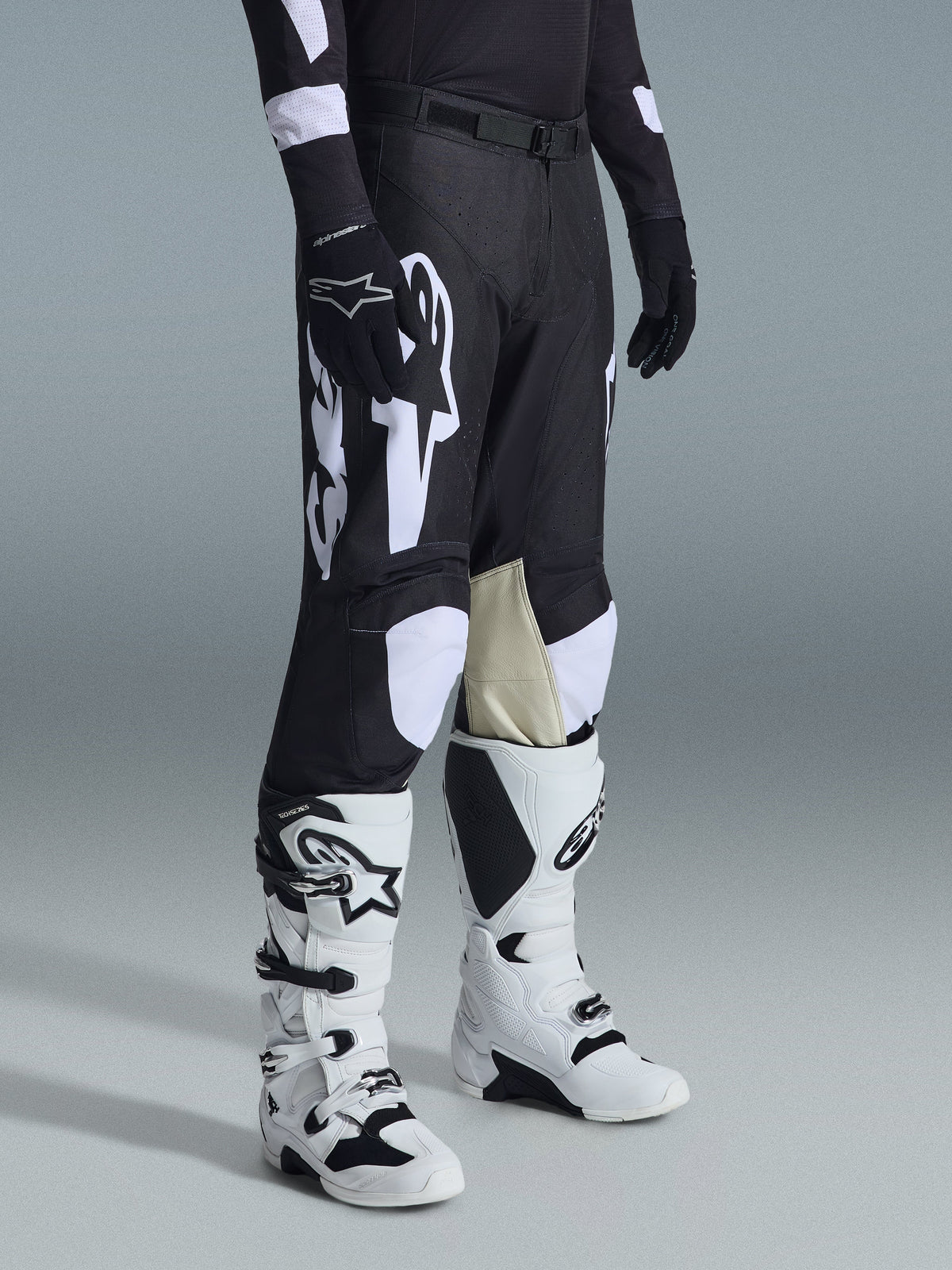Alpinestars 2026 Racer Air Riway Pants, calças de motocross em preto e branco com grandes grafismos de logótipo brancos, painéis interiores dos joelhos em pele bege, usadas com botas de todo-o-terreno brancas e pretas e luvas pretas num cenário de estúdio