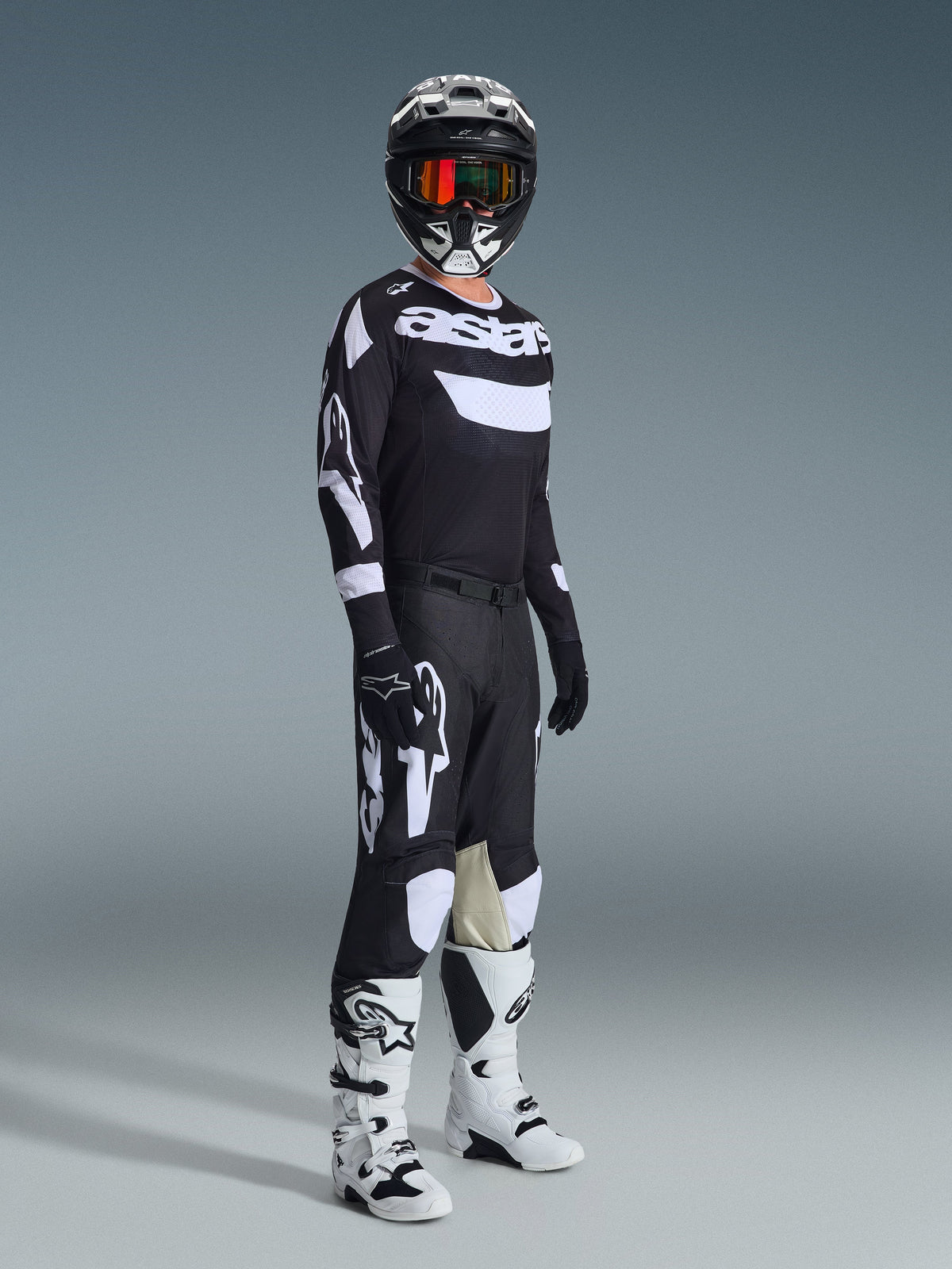 Piloto de motocross a usar Calças Racer Air Riway 2026, pretas e brancas, camisola a condizer com logótipo Alpinestars, capacete preto com óculos, botas brancas e pretas, conjunto completo de equipamento para performance em motocross todo-o-terreno