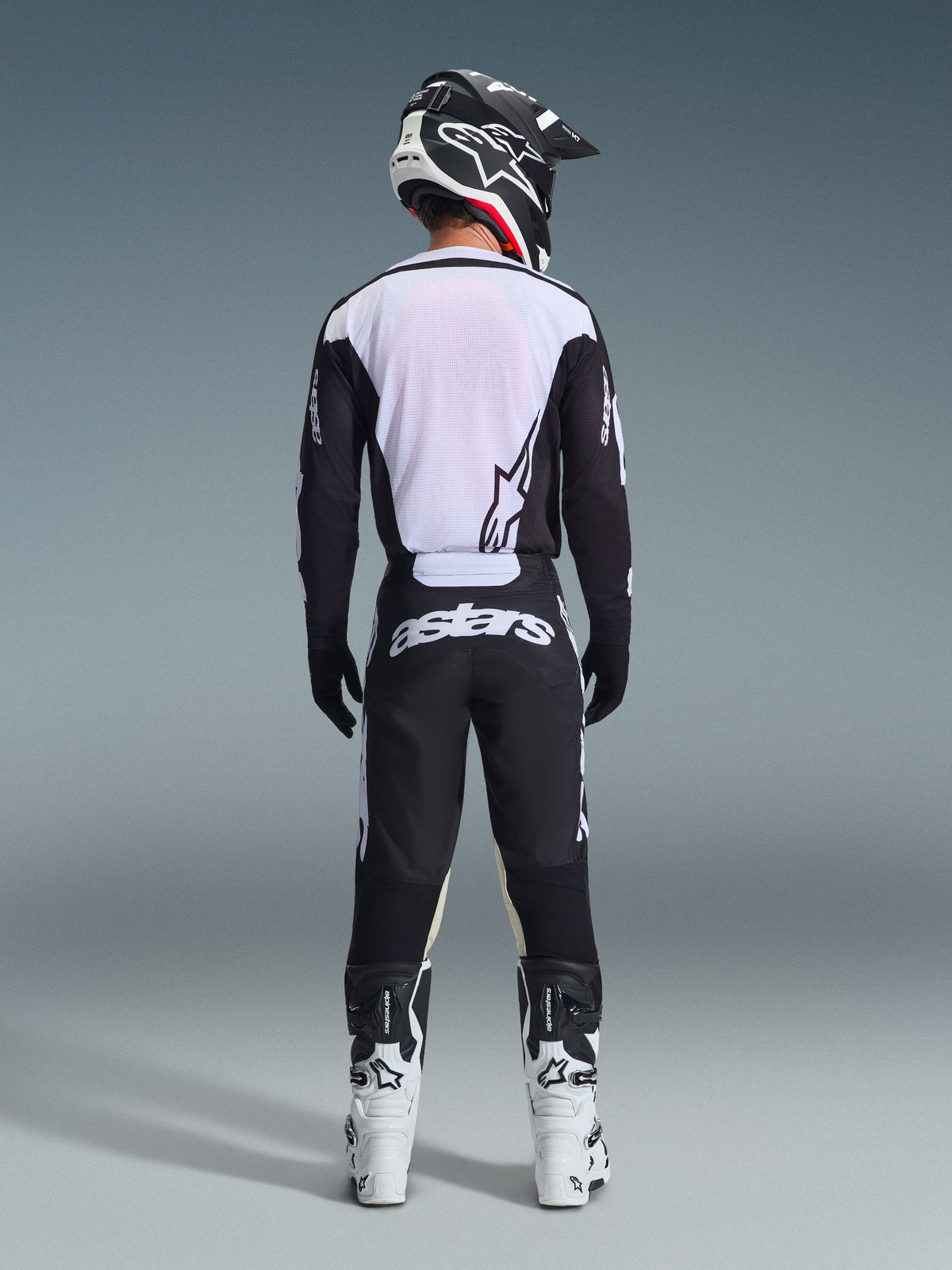 Piloto de motocross visto de costas a usar calças Alpinestars 2026 Racer Air Riway em preto e branco, camisola de manga comprida preta e branca a condizer, botas de competição brancas e pretas e capacete preto com a marca Alpinestars