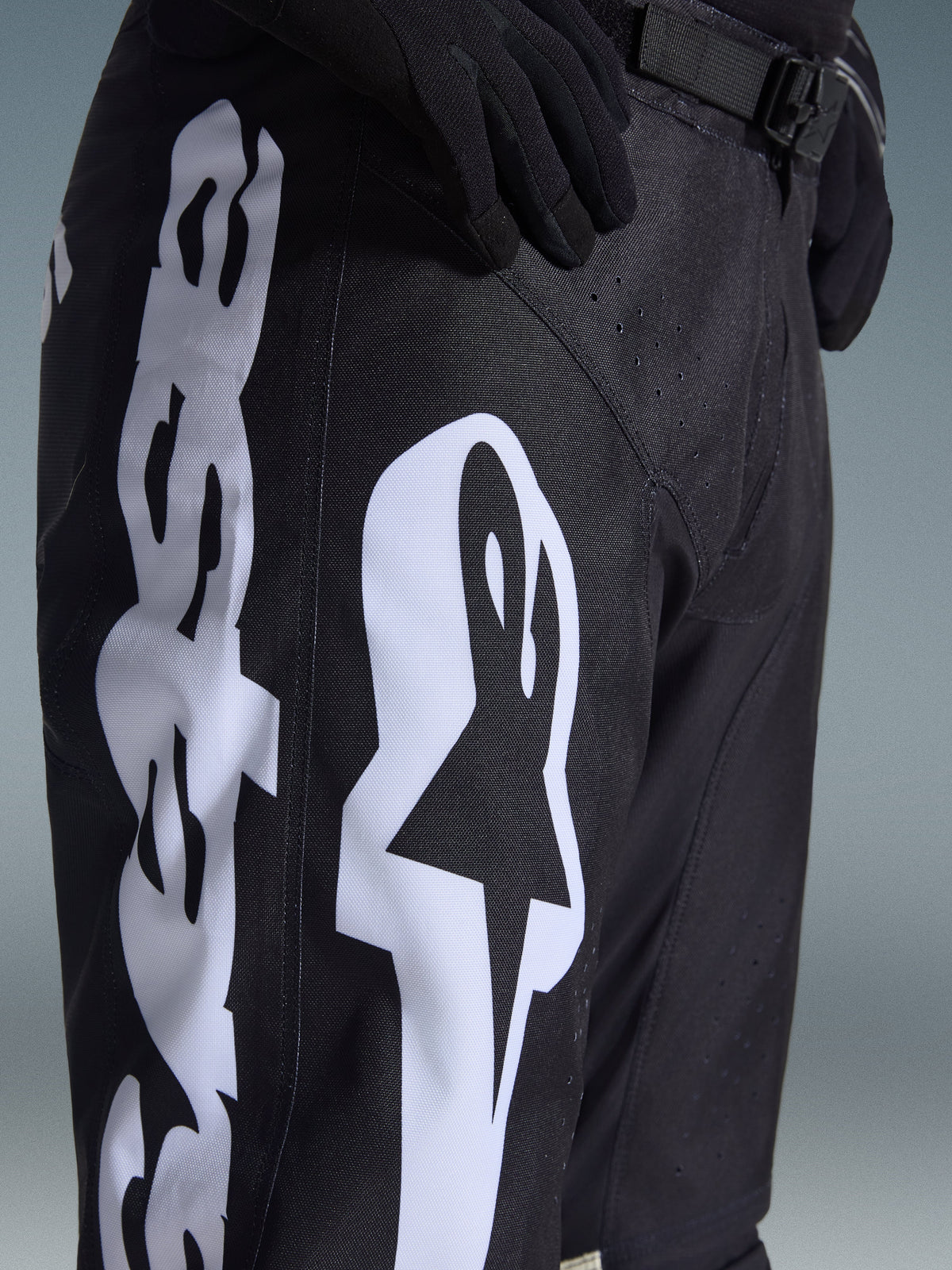 Alpinestars 2026 Racer Air Riway Pants, calças de motocross pretas com logótipo Alpinestars branco em destaque na coxa, grande plano do tecido técnico e da correia de cintura ajustável, usadas com luvas pretas para condução todo-o-terreno