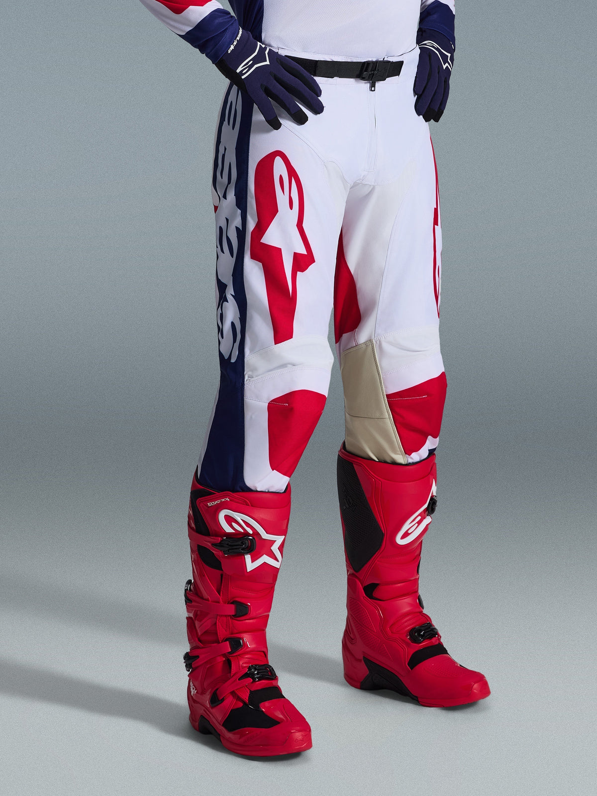 Alpinestars 2026 Racer Riway Pants, calças de motocross, branco, azul e vermelho, com logótipos vermelhos de destaque nas coxas e painéis laterais azuis com branding em branco, usadas com botas vermelhas e luvas azul-escuro, design ergonómico para corridas off-road