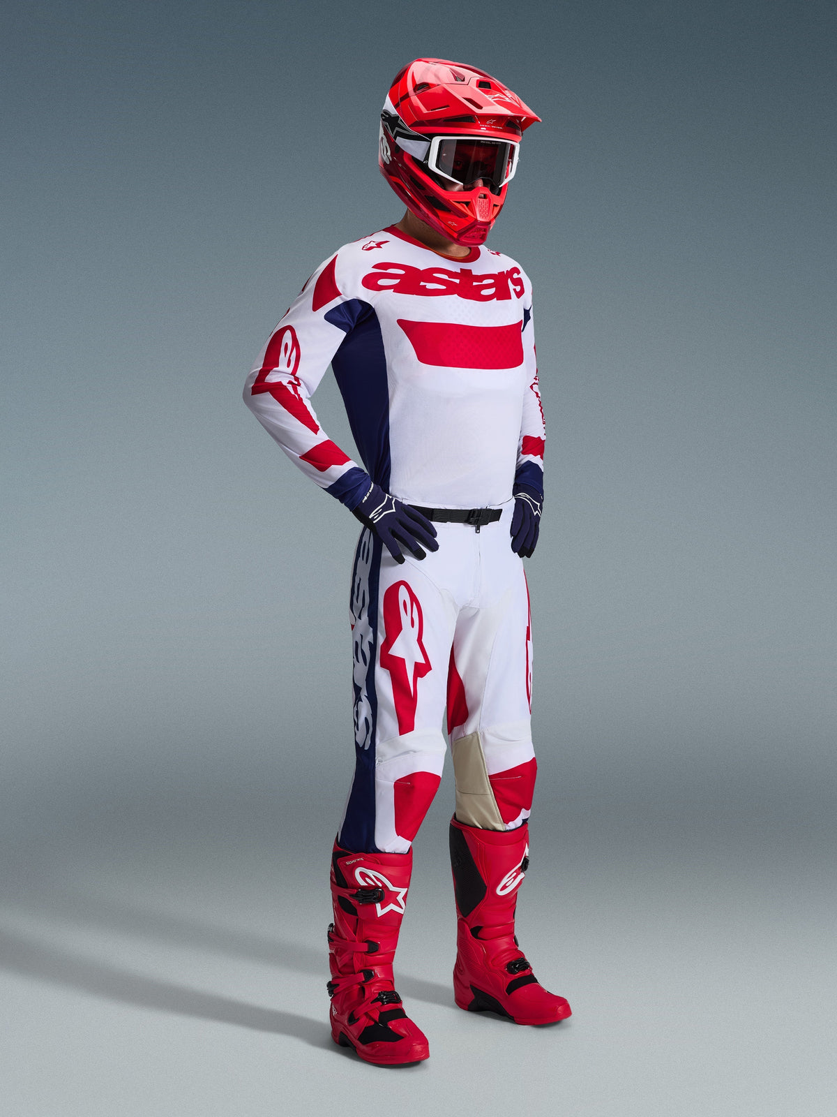 Piloto de motocross com equipamento de corrida completo, calças Alpinestars 2026 Racer Riway em branco, azul e vermelho, apresentando uma base branca com logótipos vermelhos e painéis laterais azuis, combinadas com uma camisola branca e vermelha a condizer, capacete vermelho e botas de motocross vermelhas.