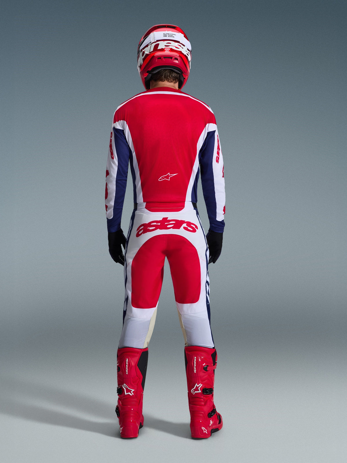 Piloto de motocross visto de costas a usar equipamento completo Alpinestars, incluindo Calças Racer Riway 2026 em branco, azul e vermelho, camisola a condizer, botas e capacete vermelhos, com logótipo 'astars' grande na parte traseira, conjunto de motocross coordenado.