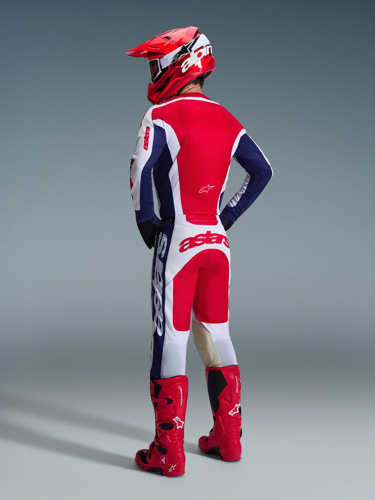 Piloto de motocross visto de costas a usar calças Alpinestars 2026 Racer Riway em branco, azul e vermelho, combinadas com uma camisola a condizer, capacete vermelho e botas vermelhas vibrantes, exibindo o logótipo arrojado no assento e o ajuste atlético de todo-o-terreno.