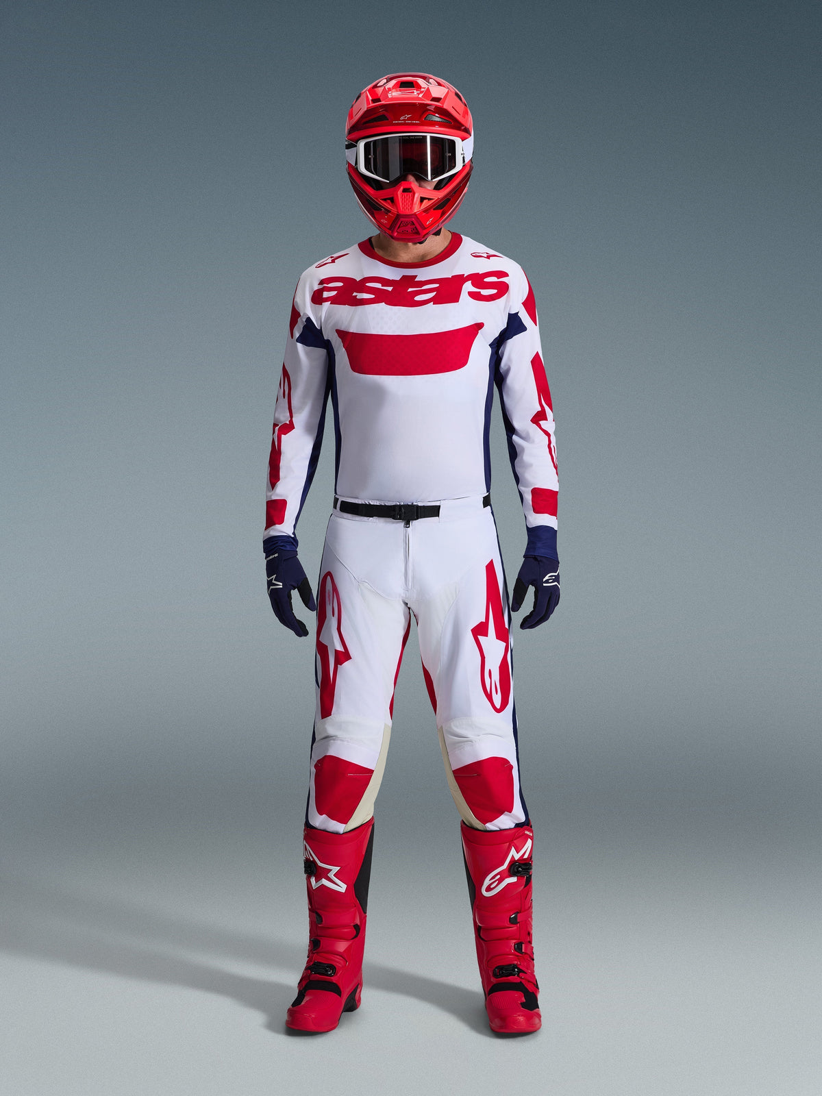 Piloto de motocross com equipamento de corrida completo, camisola branca com marca Alpinestars a vermelho e pormenores em azul, Calças Alpinestars 2026 Racer Riway em Branco Azul Vermelho, capacete vermelho com óculos a condizer, botas vermelhas e pretas, luvas em azul-escuro, conjunto coordenado para corridas todo-o-terreno