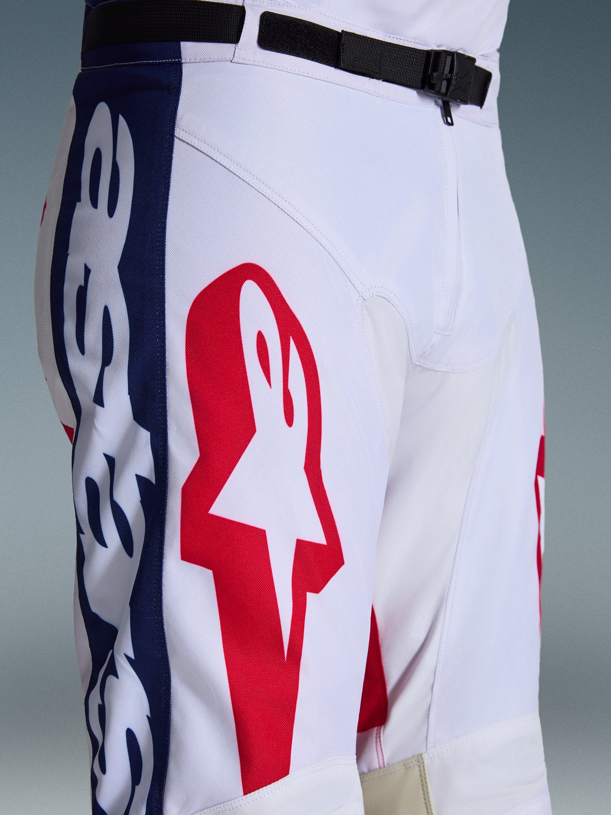 Calças Alpinestars Racer Riway 2026, calças de motocross, brancas, azuis e vermelhas, detalhe da parte superior da perna e cintura com grande logótipo de estrela vermelha em tecido poliéster branco, painéis laterais azuis com marca, cinto ajustável preto com fivela magnética SX2, design desportivo de corrida todo-o-terreno