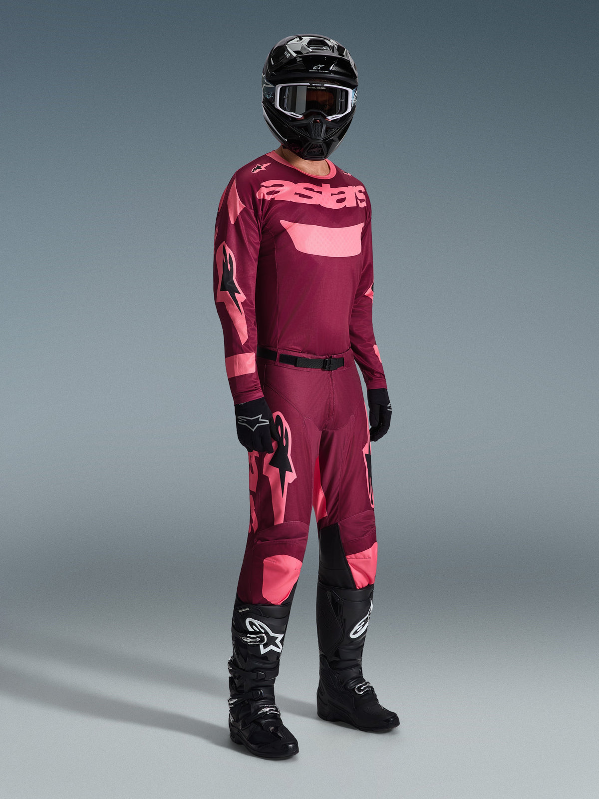 Piloto de motocross com equipamento de competição completo, Camisola Alpinestars Racer Riway 2026 em Dark Red Coral com detalhes rosa, calças vermelho escuro a condizer com logótipos rosa, capacete preto com óculos de moldura branca, luvas pretas e botas pretas com logótipos brancos, conjunto coordenado para corridas todo-o-terreno