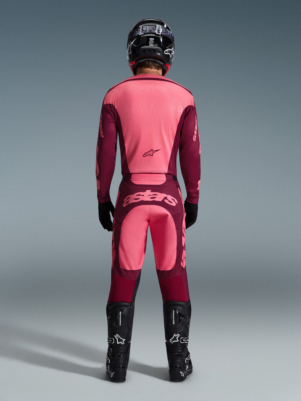 Piloto de motocross visto de costas a usar a camisola Alpinestars 2026 Racer Riway e calças a condizer em Dark Red Coral, com botas pretas, capacete preto e luvas pretas, equipamento de alto desempenho coordenado para todo-o-terreno
