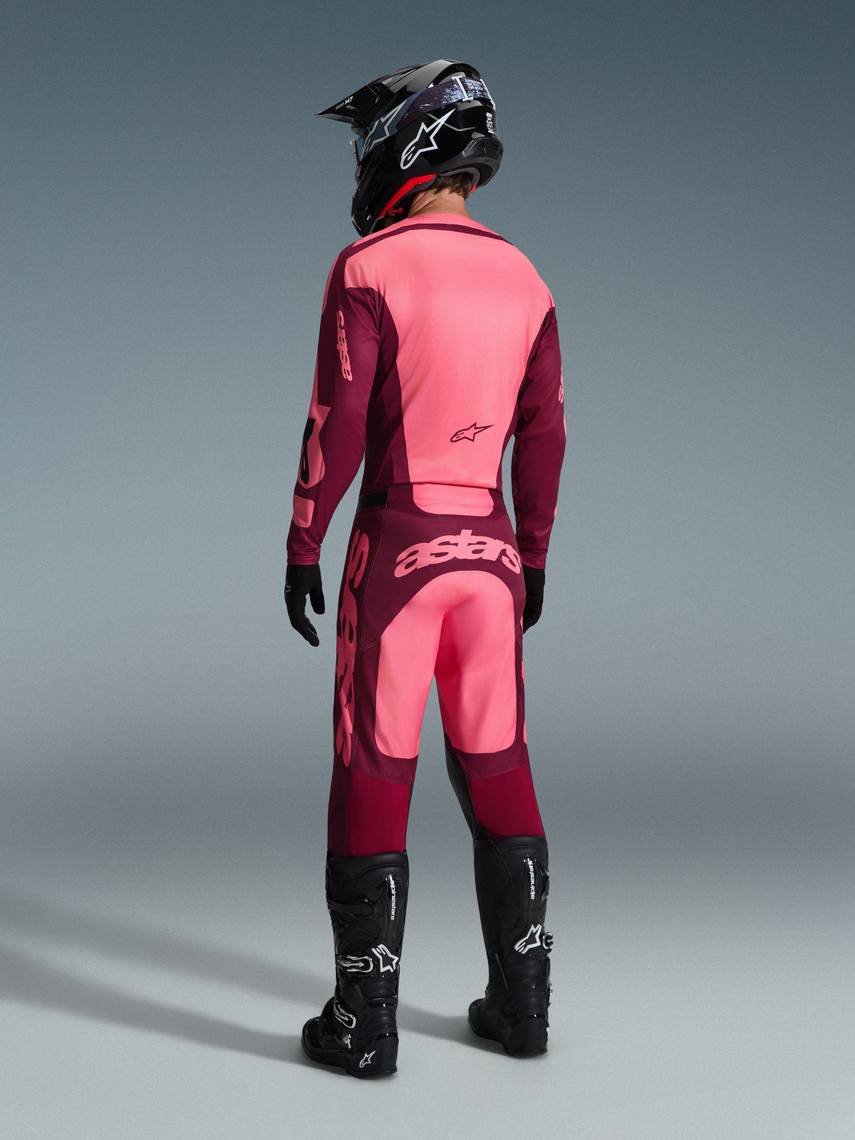 Camisola Alpinestars 2026 Racer Riway e calças a condizer, equipamento de motocross em Dark Red Coral, vista traseira do piloto com capacete preto e botas pretas, apresentando painéis vibrantes em coral e bordeaux com branding Astars em destaque nas costas