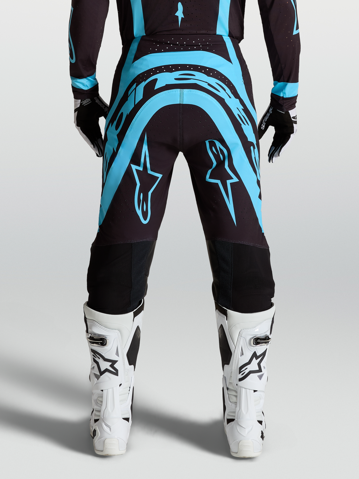 Alpinestars Calças Supertech Vesta, calças de motocross para homem, preto e turquesa com botas todo-o-terreno brancas, vista traseira, ajuste de corrida racing fit