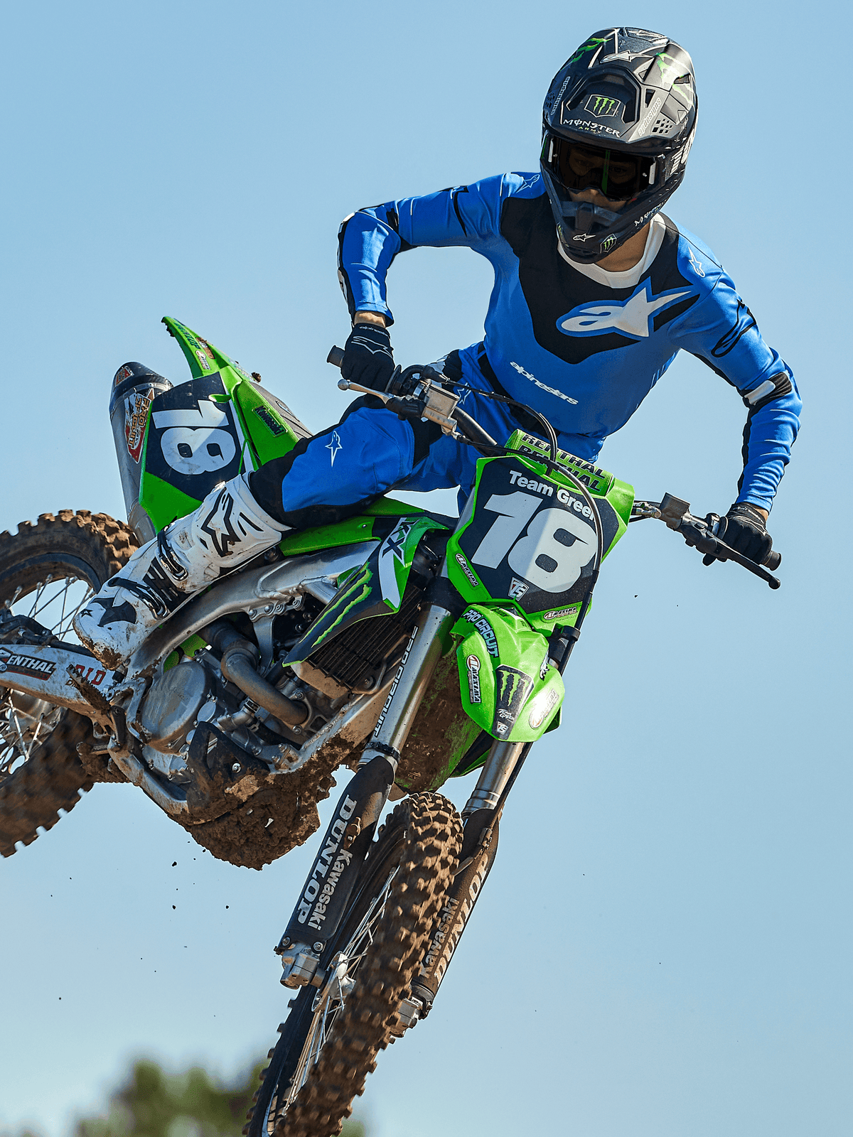 Piloto de motocross em pleno salto numa mota verde, com calças Alpinestars 2025 Racer Veil em azul e preto, camisola azul com logótipo branco no peito, botas brancas e pretas e capacete preto, equipamento de alto desempenho para competição em todo-o-terreno.