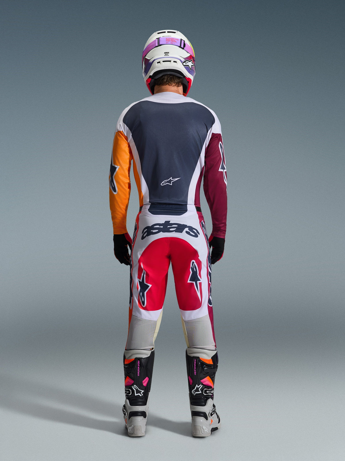 Piloto de motocross visto de costas a usar as calças Alpinestars Racer Portl 2026 em vermelho, cinzento claro e cinzento escuro, com a marca "astars" no assento, combinadas com uma camisola multicolorida, capacete branco e botas de competição técnicas.