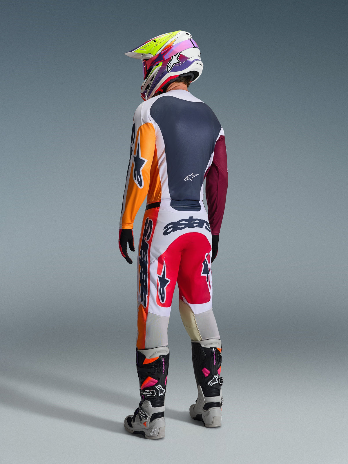 Piloto de motocross visto de costas a usar equipamento de corrida completo, calças Alpinestars 2026 Racer Portl em Vermelho, Cinzento Claro e Cinzento Escuro, camisola a condizer com mangas cor de laranja e bordeaux, capacete com grafismos multicoloridos, botas de motocross cinzentas e pretas com detalhes cor-de-rosa, conjunto de motocross profissional