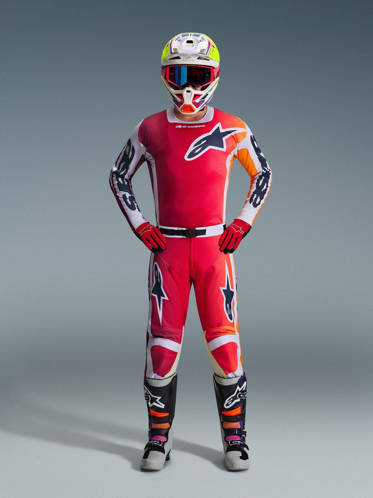 Piloto de motocross Alpinestars com equipamento completo, camisola vermelha com detalhes em branco e laranja, calças Racer Portl 2026 em vermelho e branco, capacete branco com detalhes coloridos e óculos azuis, luvas vermelhas e botas pretas e cinzentas, conjunto coordenado para desempenho off-road
