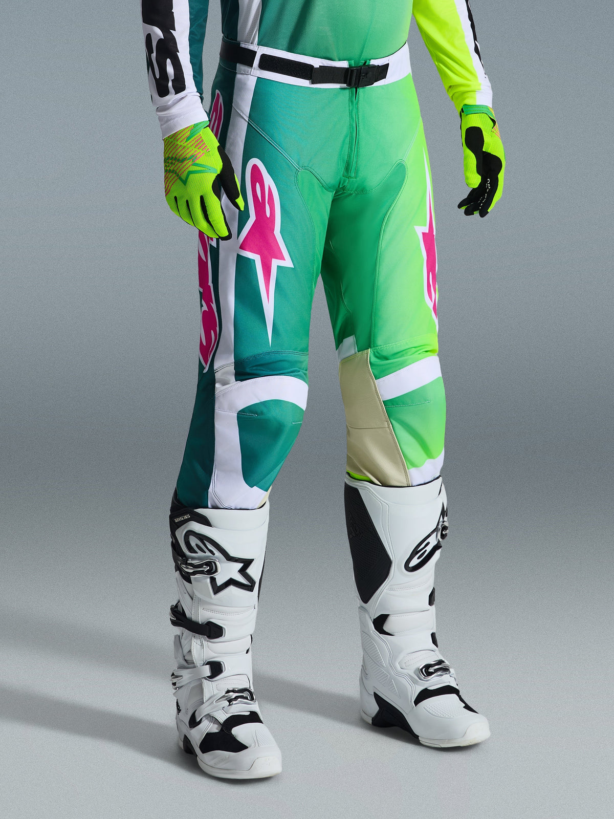 Alpinestars 2026 Racer Portl Pants, calças de motocross, verdes, brancas e pretas com logótipos cor-de-rosa, usadas com botas brancas e luvas amarelo néon, com padrões gráficos arrojados e uma fivela de cintura preta ajustável, equipamento de performance para condução todo-o-terreno