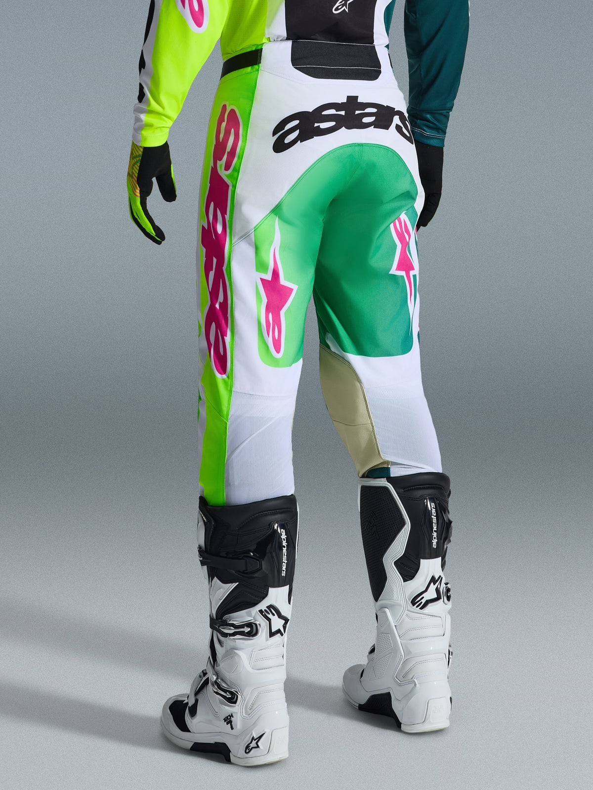 Alpinestars 2026 Racer Portl Pants, calças de motocross, verde, branco e preto com detalhes cor-de-rosa, vista traseira apresentando o logótipo 'astars' em destaque no assento e logótipos de estrela cor-de-rosa nas pernas, combinadas com botas de motocross brancas e pretas, equipamento de performance para corridas off-road
