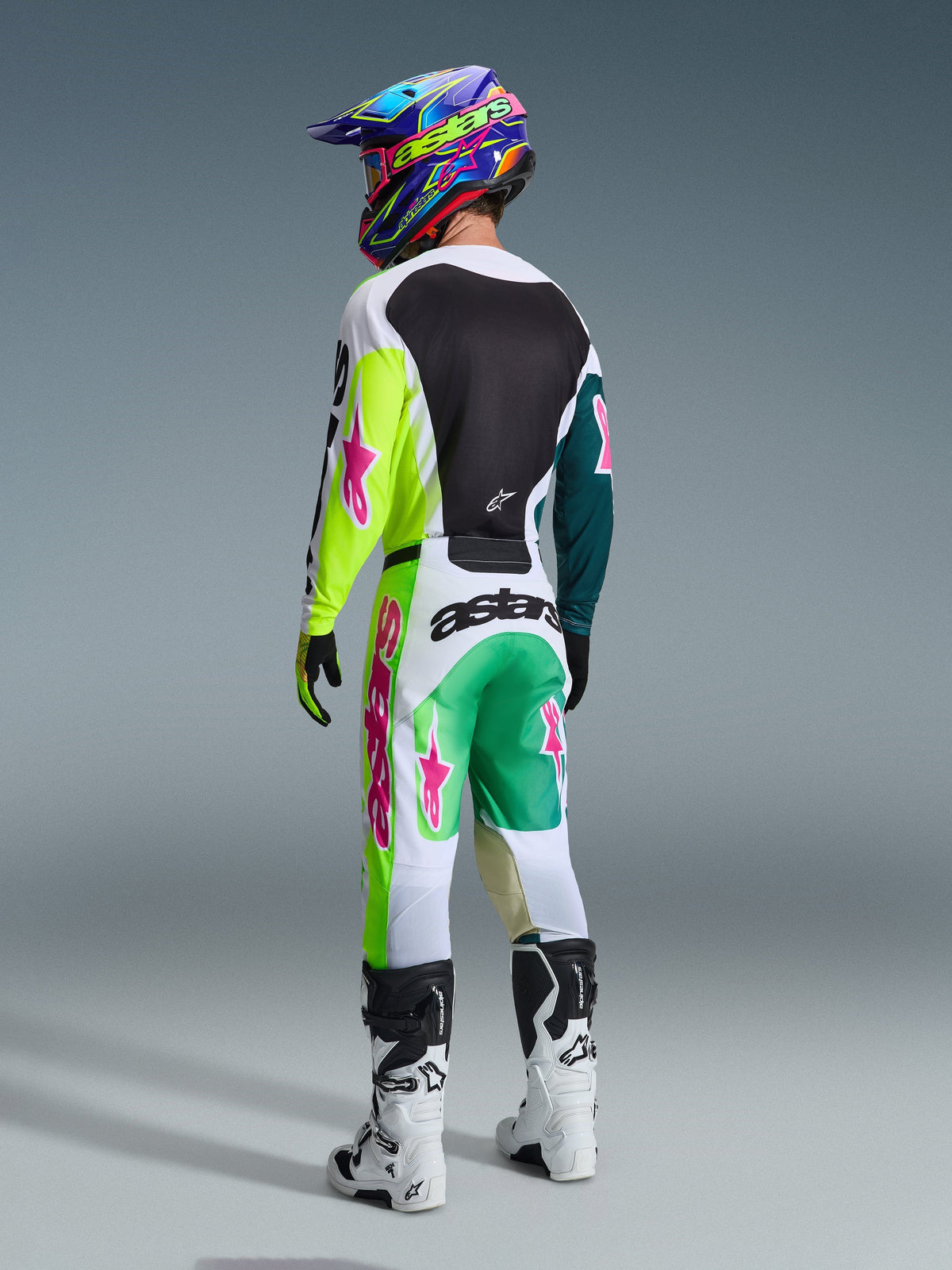 Piloto de motocross visto de costas a usar as calças Alpinestars 2026 Racer Portl em verde, branco e preto, camisola a condizer com detalhes em lima e rosa, capacete colorido, botas brancas e pretas, conjunto completo de equipamento de corrida off-road.