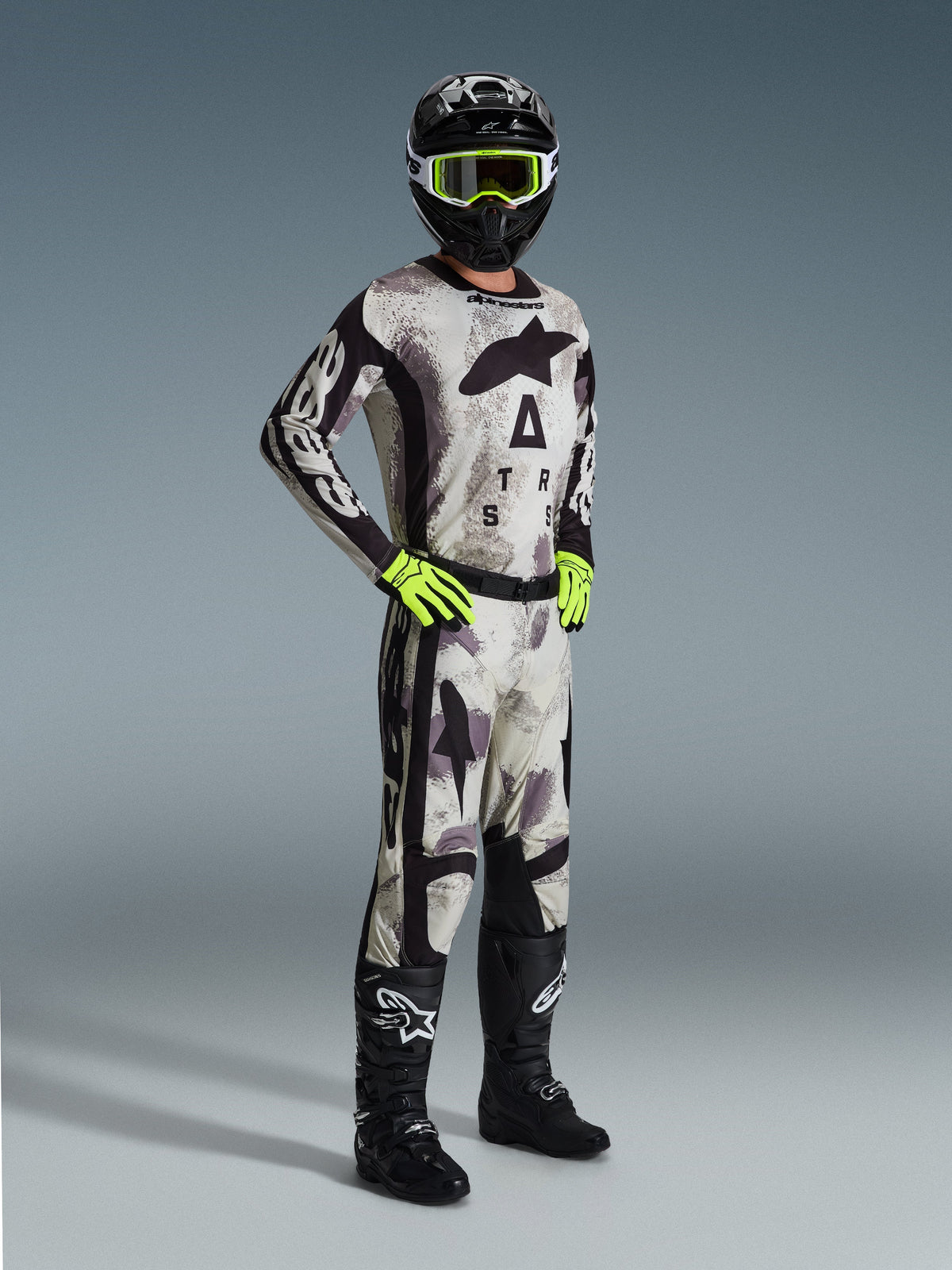 Piloto de motocross a usar a Camisola Alpinestars Racer Lahnd 2026 em Sand Camo, com padrão de camuflagem cinzento e preto, calças de motocross a condizer, luvas amarelo néon, capacete preto com óculos amarelos e botas pretas, equipamento profissional completo para corridas off-road.
