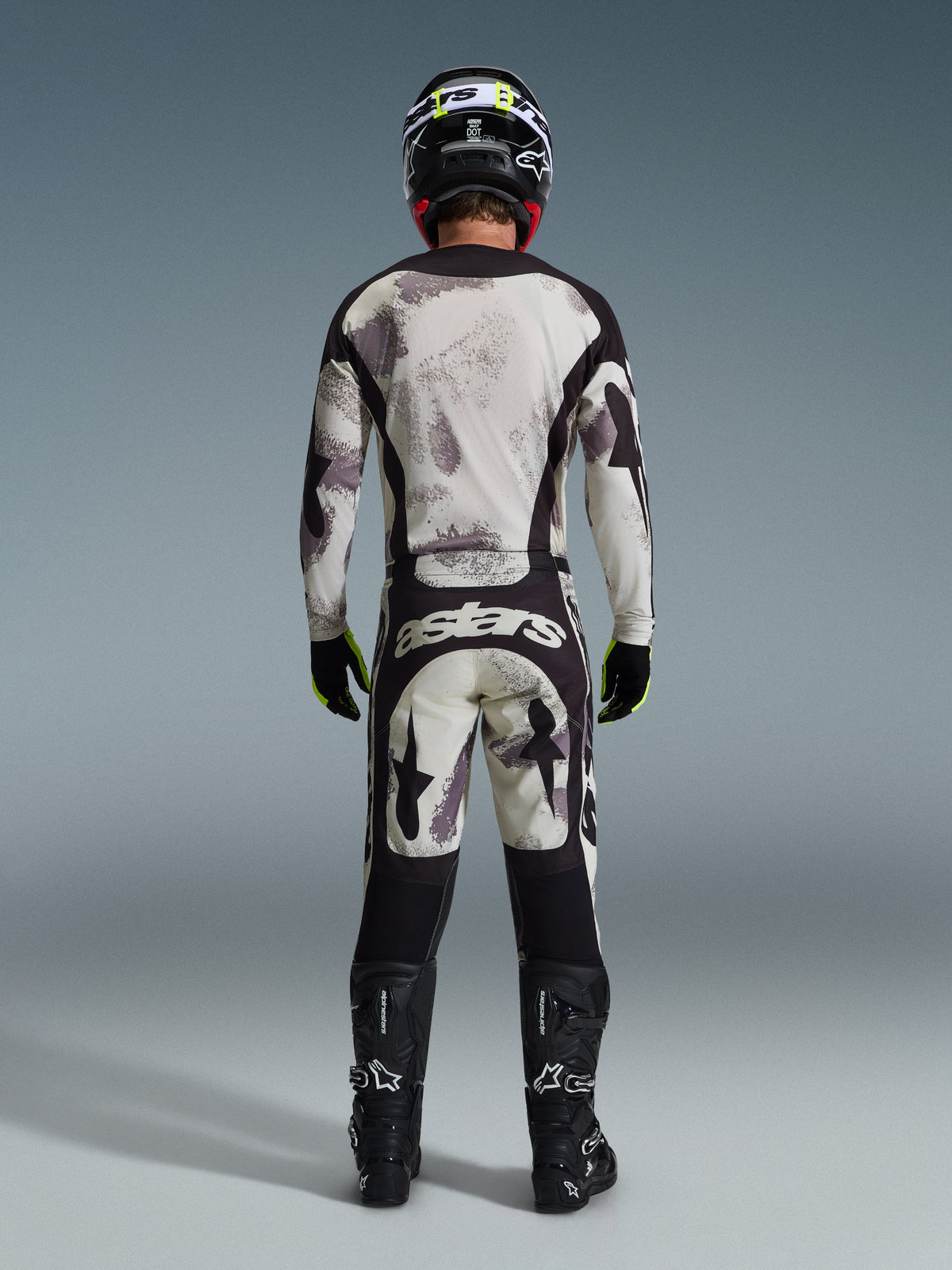 Piloto de motocross visto de costas a usar a Camisola Racer Lahnd 2026 e calças a condizer em Sand Camo, com um padrão abstrato creme e cinzento e painéis pretos, marca 'astars' nas calças, em conjunto com botas Alpinestars pretas e um capacete preto para um visual de corrida off-road completo.
